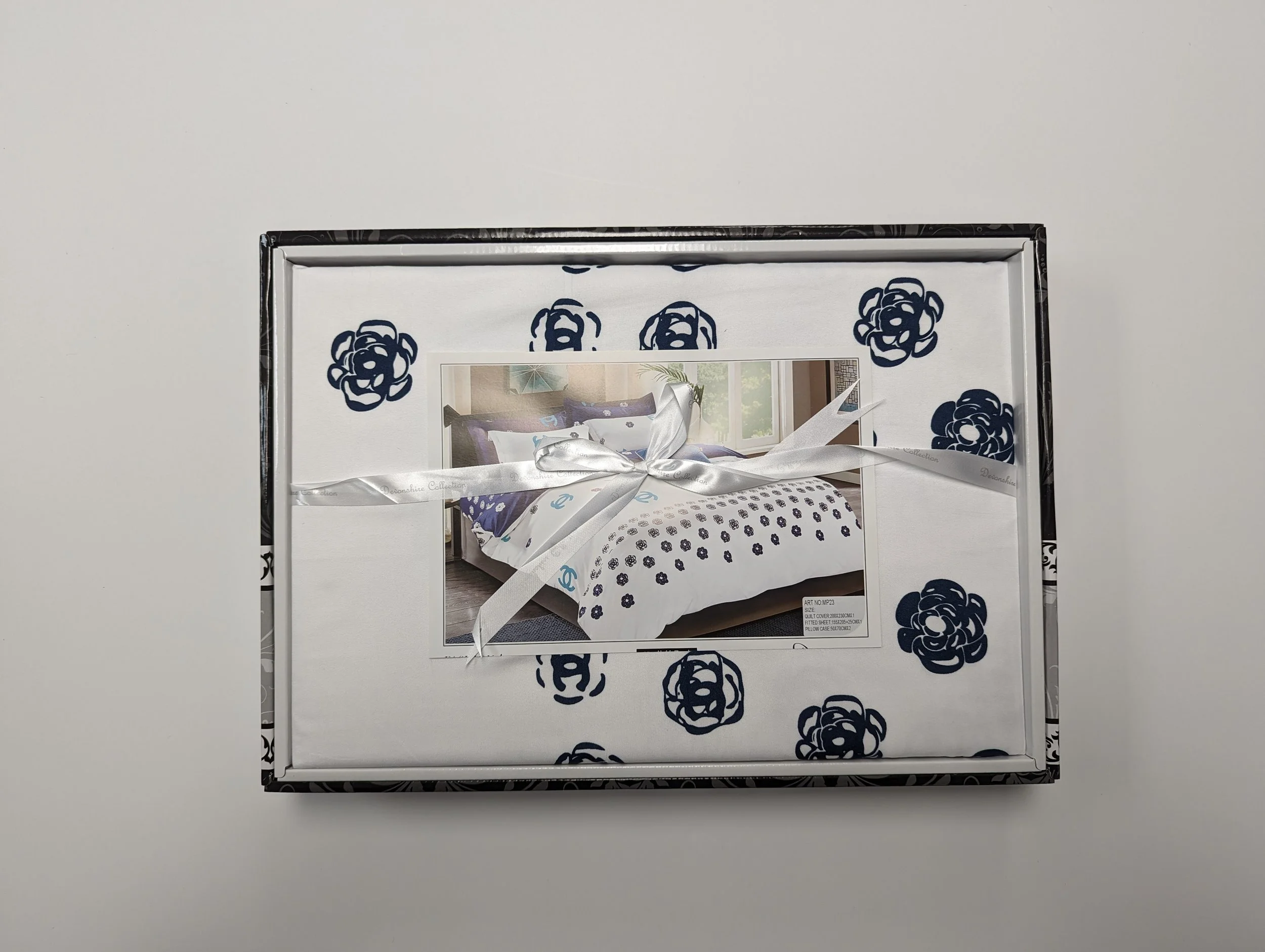 Box containing bedding with a photo of a bed inside, decorated with navy floral patterns, white ribbon tying the box
