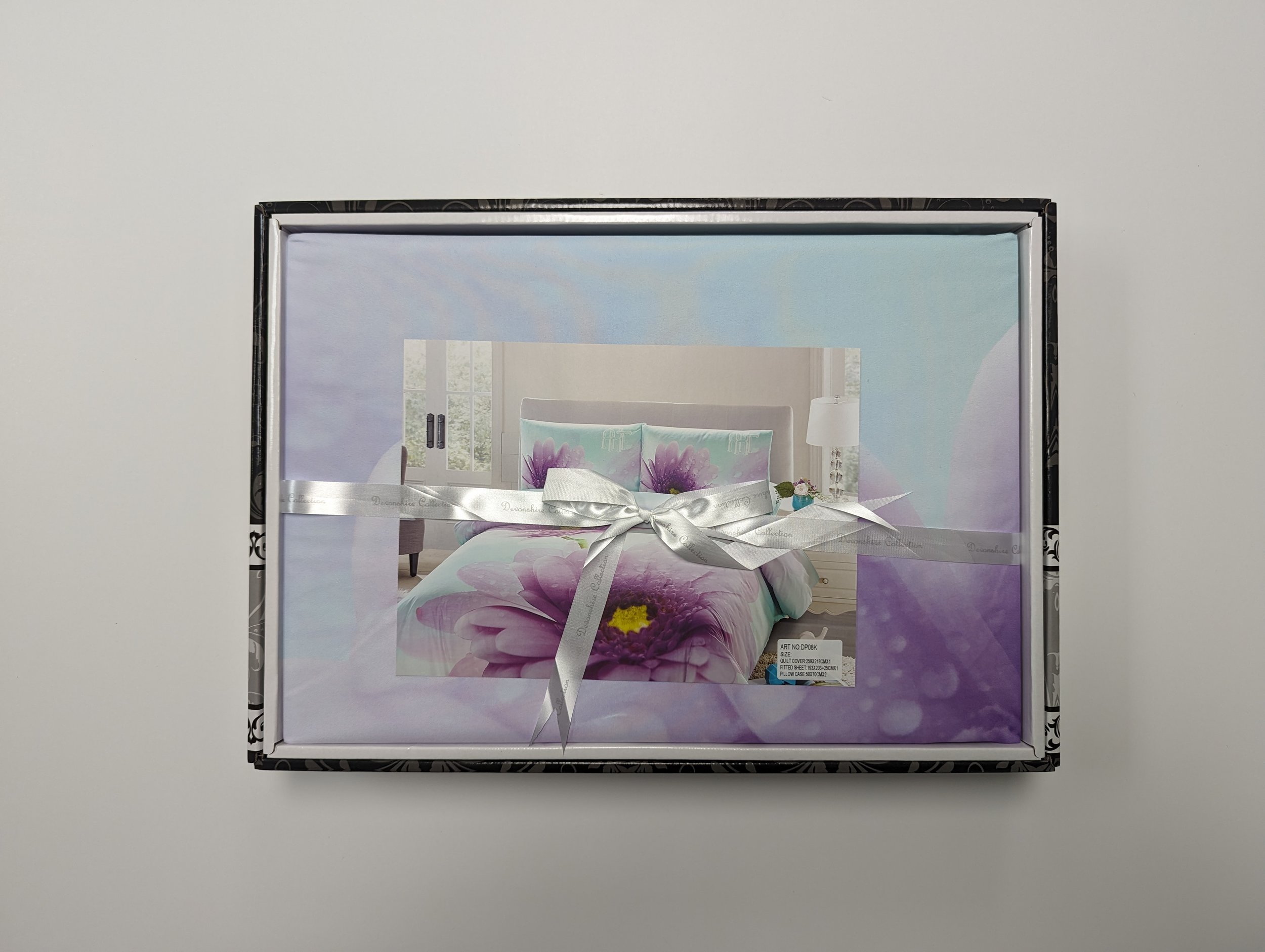 Box with floral-themed bedding set featuring purple and pink flowers, wrapped with a white ribbon.