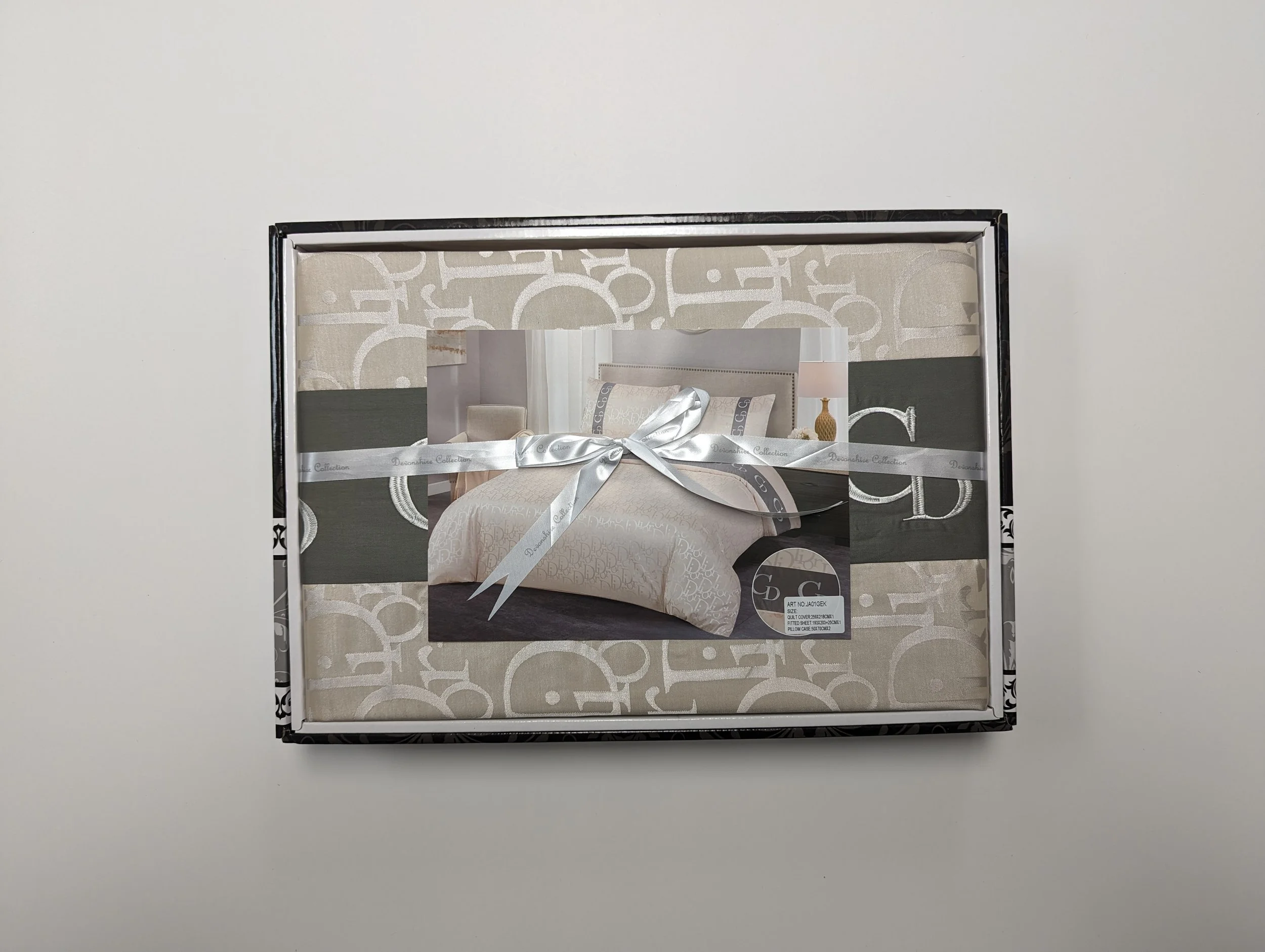 Box containing a beige and white bedding set with pillows and comforter, wrapped with a white satin ribbon.