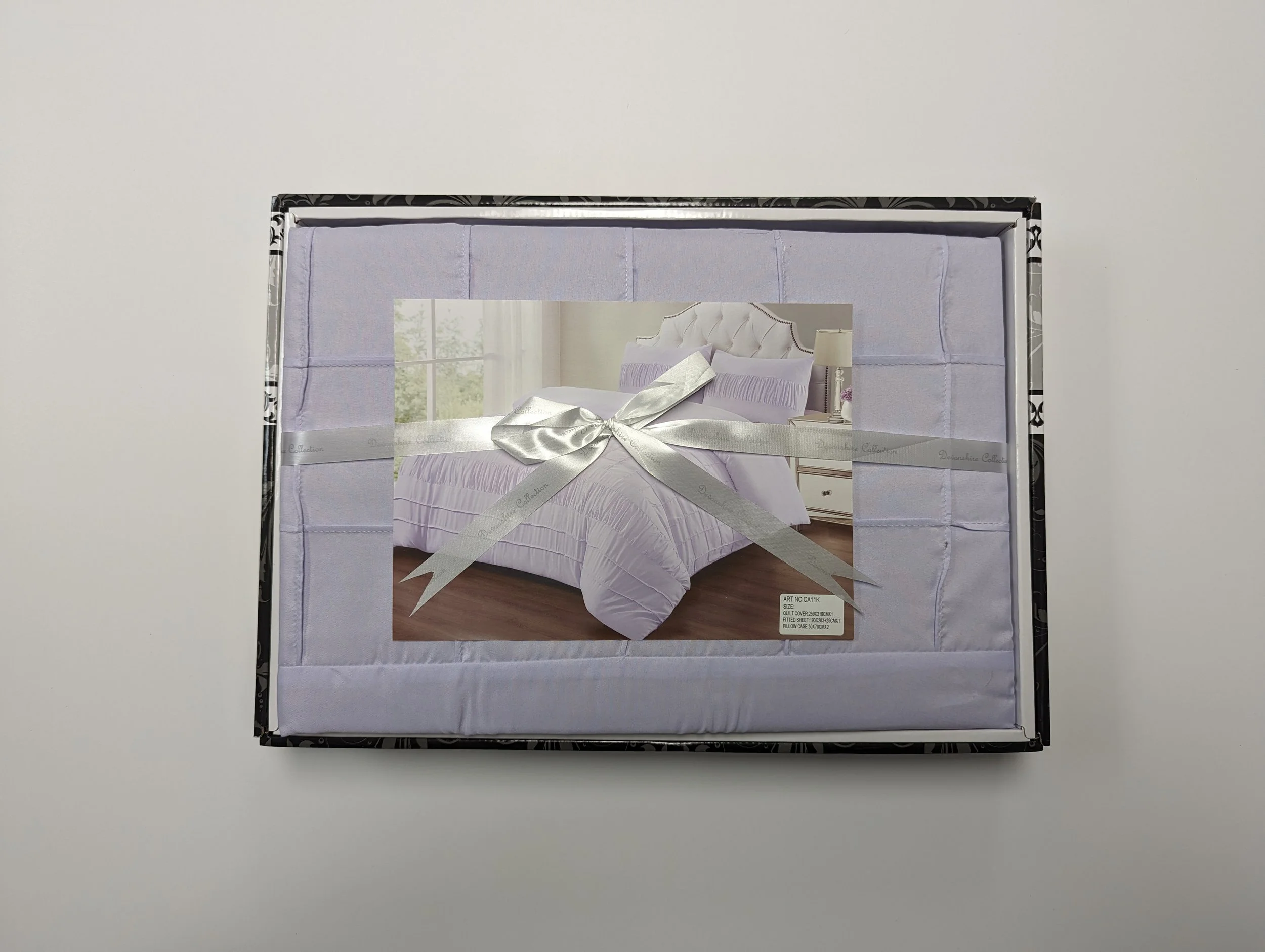 A boxed set of purple bed linens with a picture of a bedroom with a bed and nightstand, wrapped with a silver ribbon.