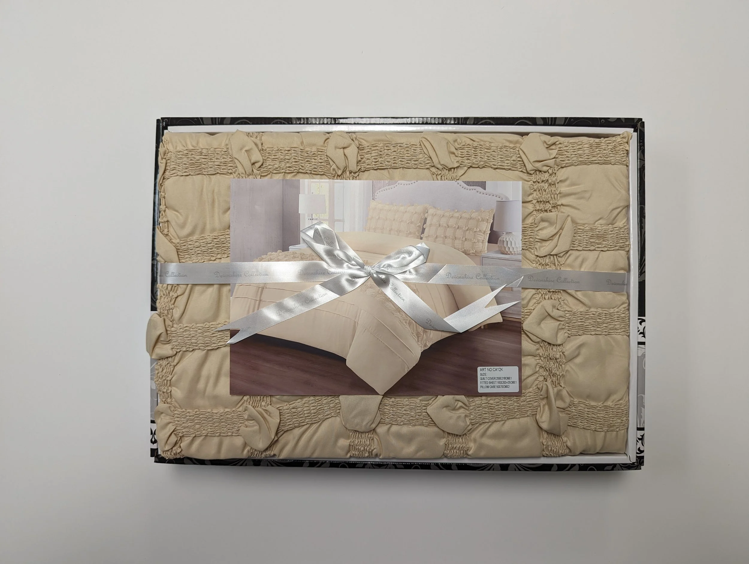 Packaging of a beige bedspread with textured accents, including a photo of a bedroom with matching bed and pillows attached with a silver ribbon.