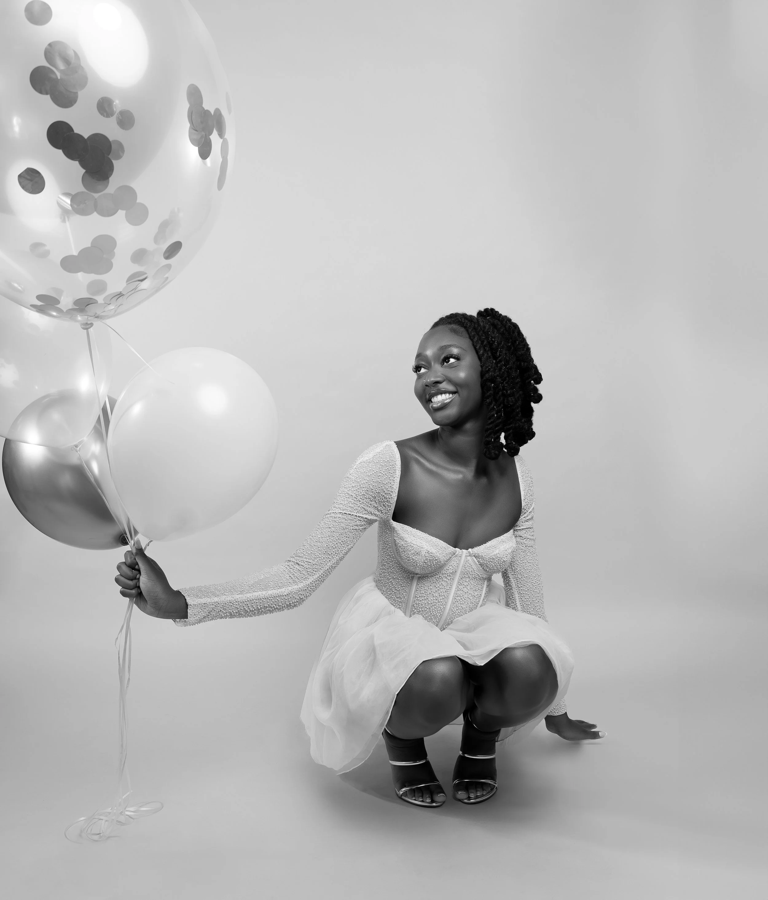 Smiling woman in a white dress holding balloons in a studio setting.