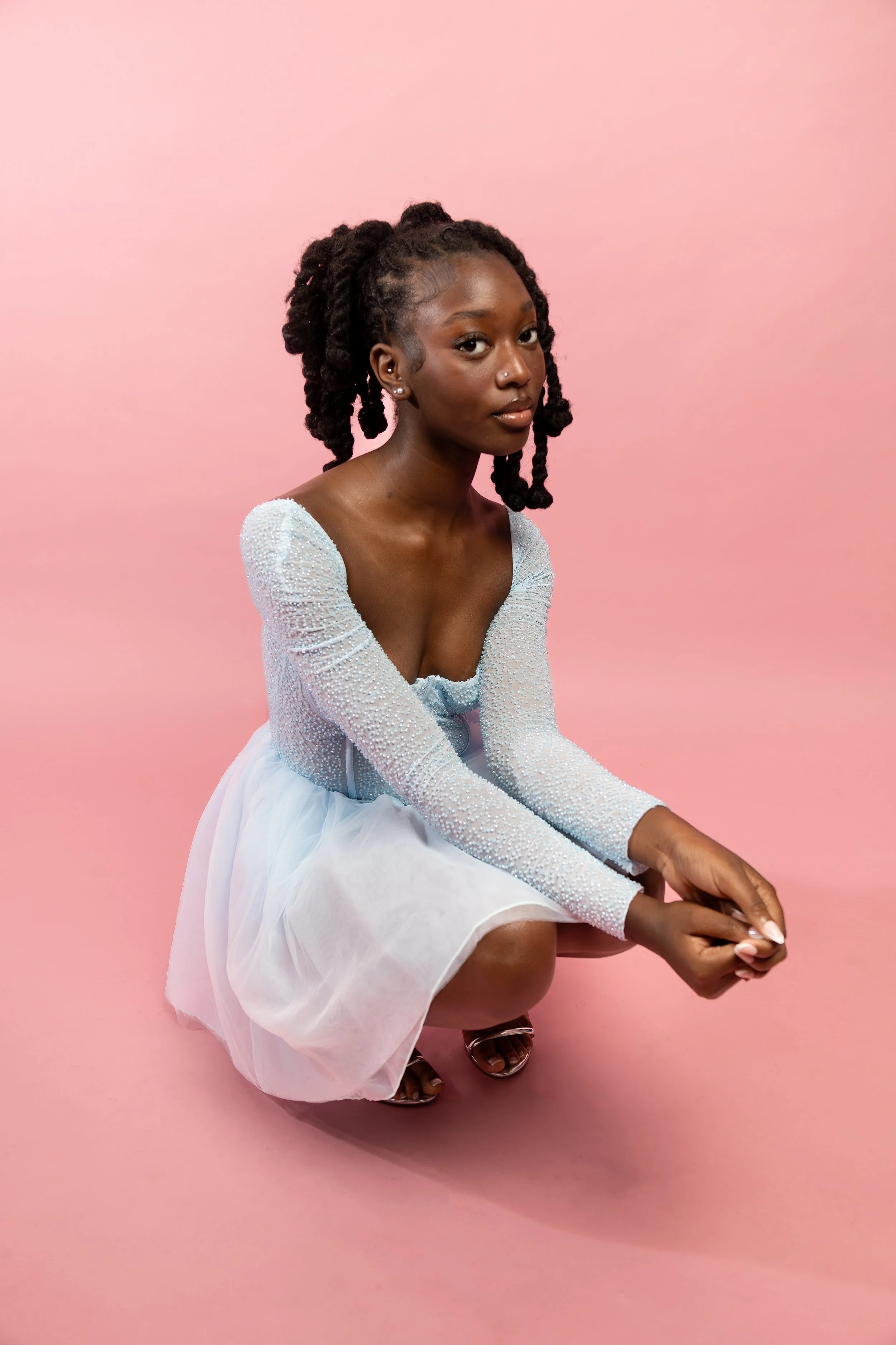 Young woman in light blue dress with textured sleeves kneeling on pink background.