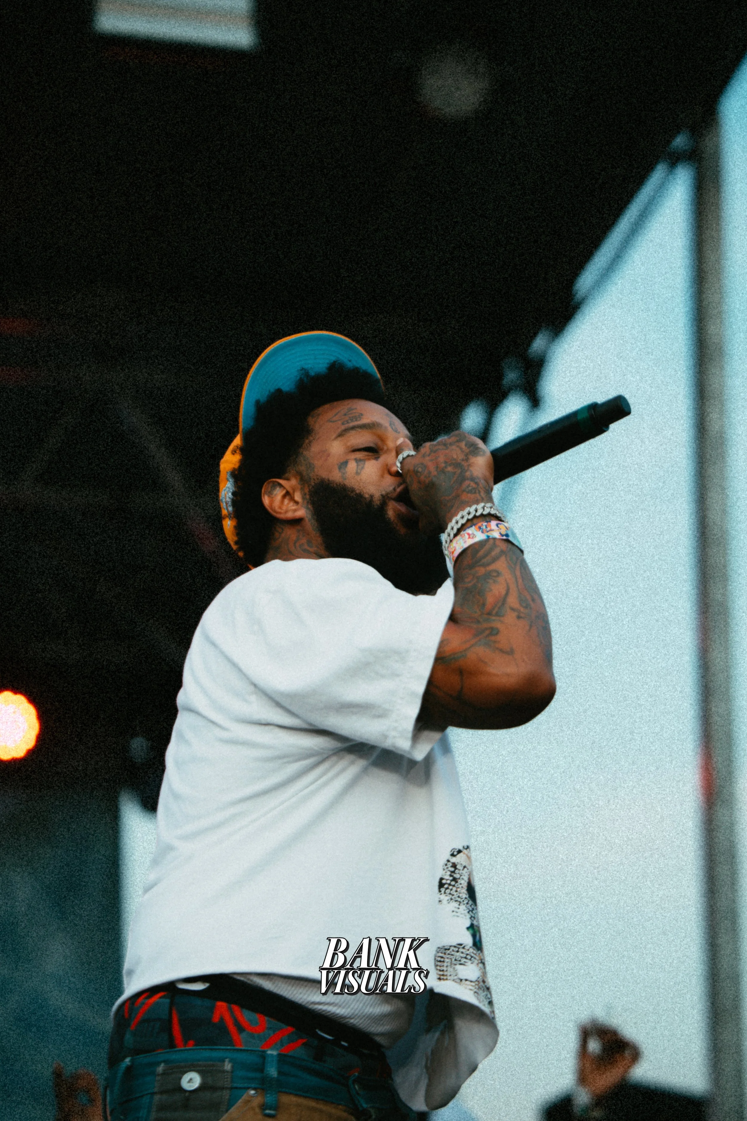 Performer with microphone on stage, wearing a white shirt and hat, tattoos visible.