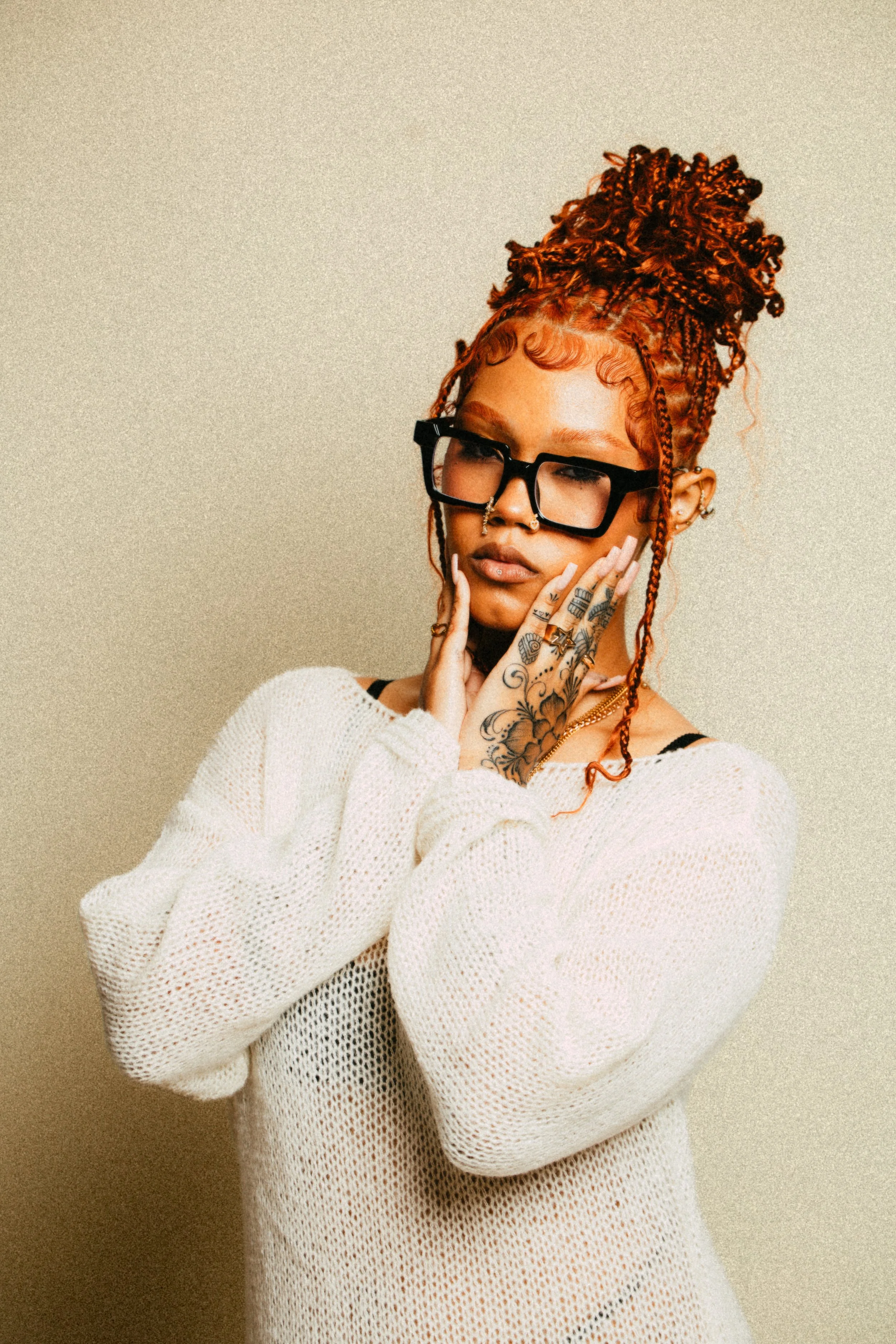 Person with red curly hair in a top bun, wearing glasses, a white sweater, and featuring detailed hand tattoos, posing against a plain background.