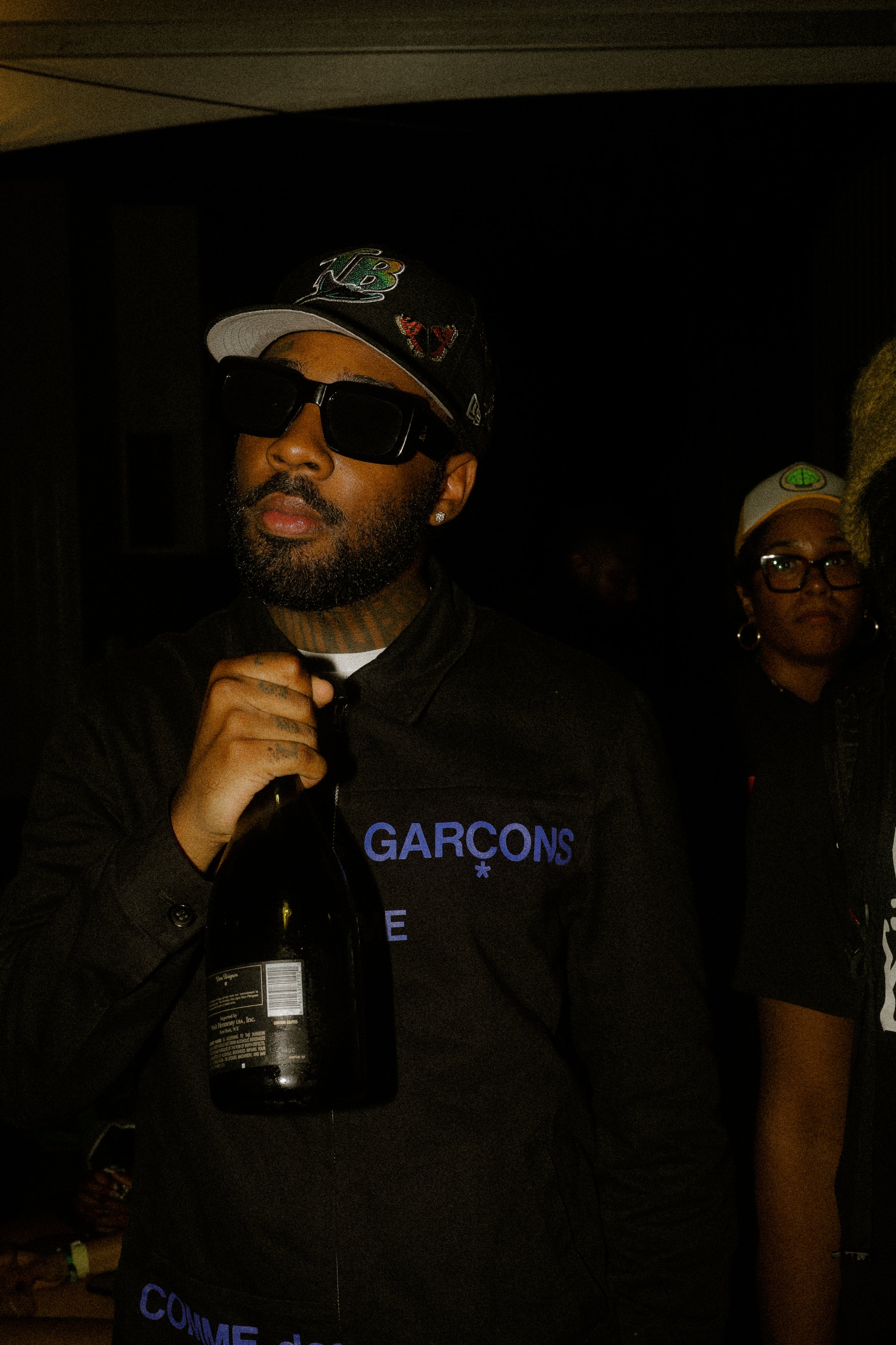 Person wearing a cap and sunglasses holding a bottle, with another person in the background wearing glasses and a cap.