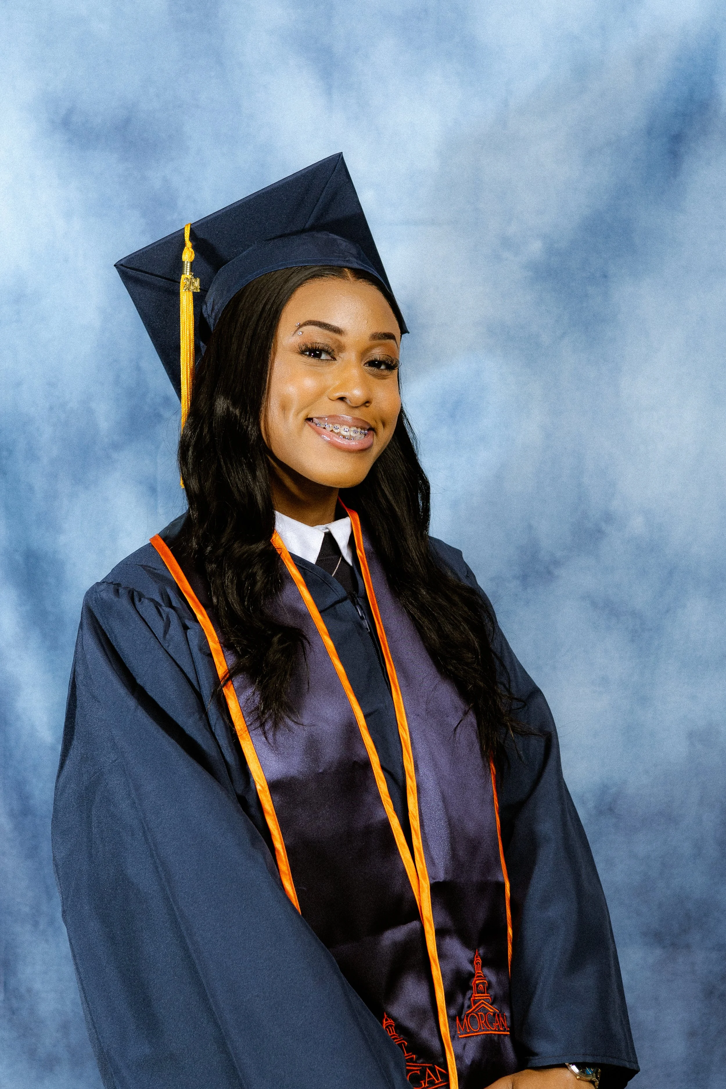 Graduate wearing a blue cap and gown with orange accents, smiling against a blue background.