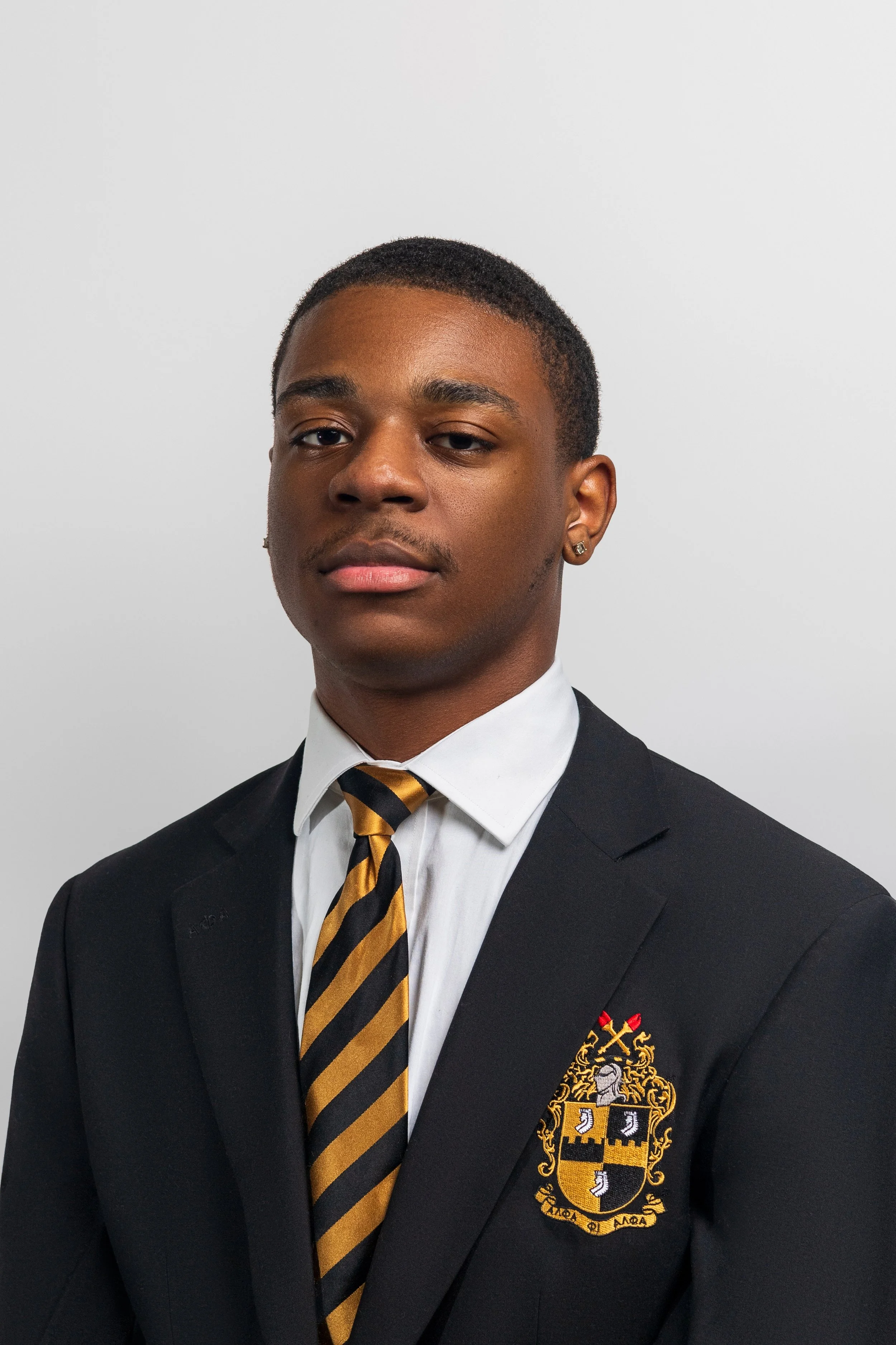 Young man in a suit with a striped tie and crest, standing against a plain background.