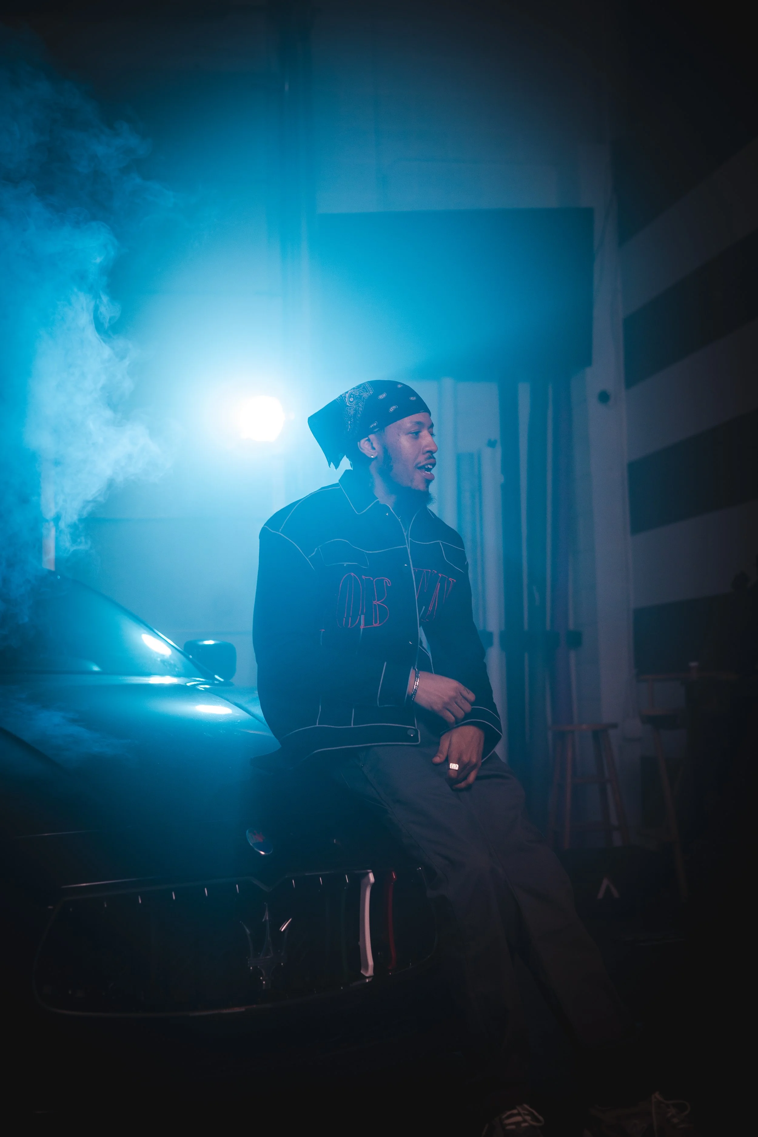 Man in dark clothing and bandana sitting on a car hood with blue atmospheric lighting and smoke.