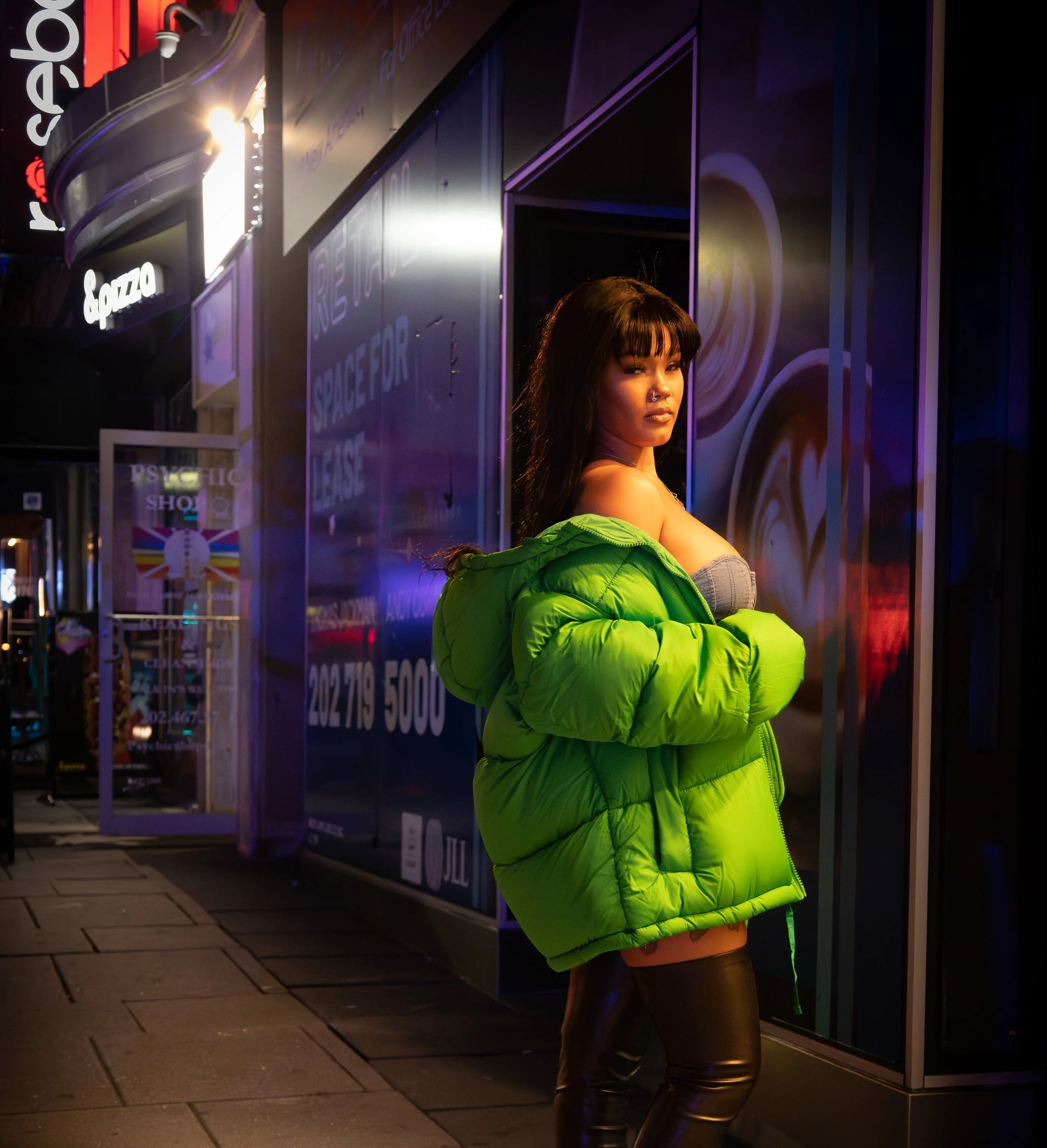 Woman in a bright green jacket posing on a city street at night with neon lights.