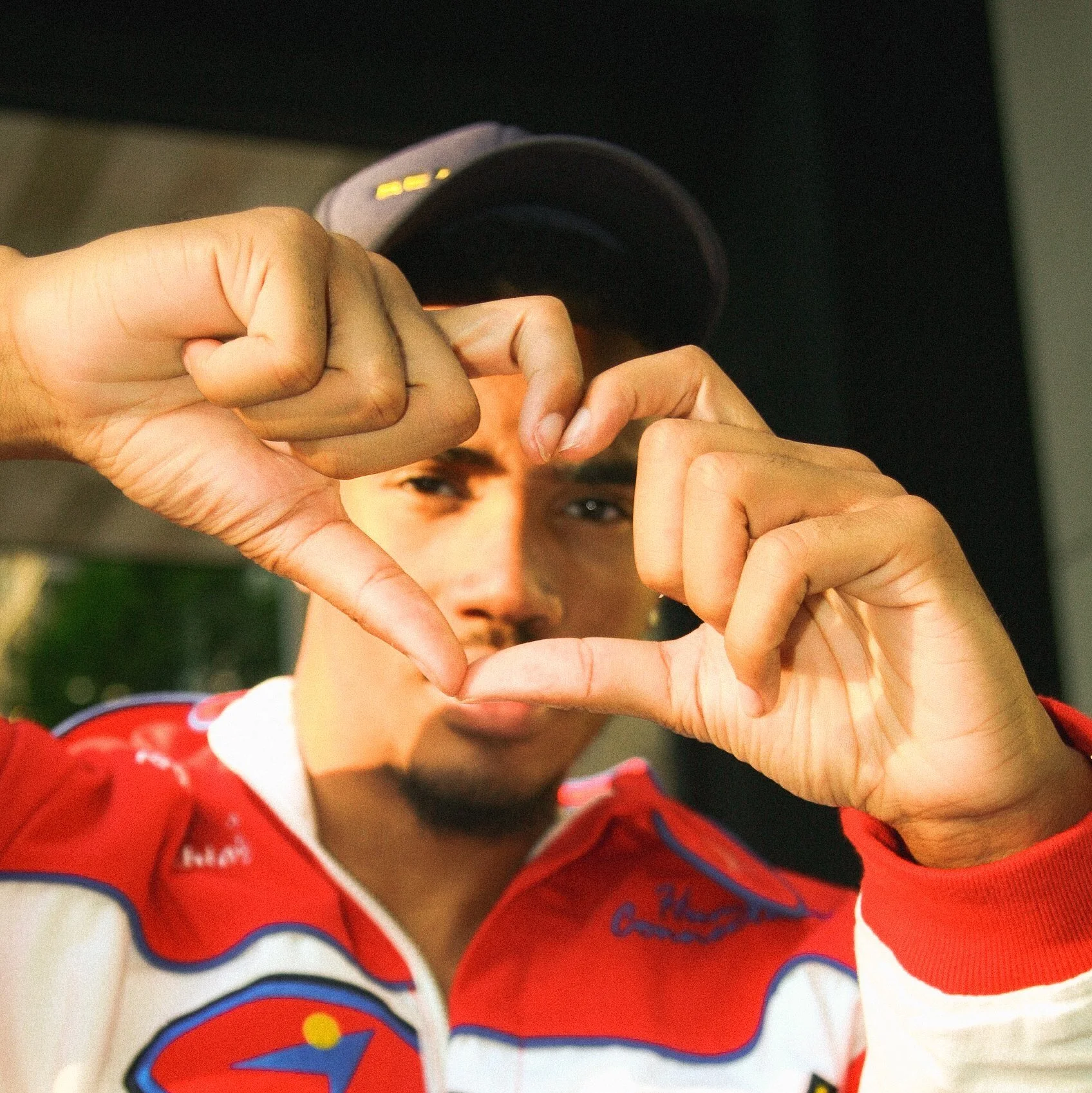 Person making a heart shape with hands, wearing a cap and red jacket with white and blue details.
