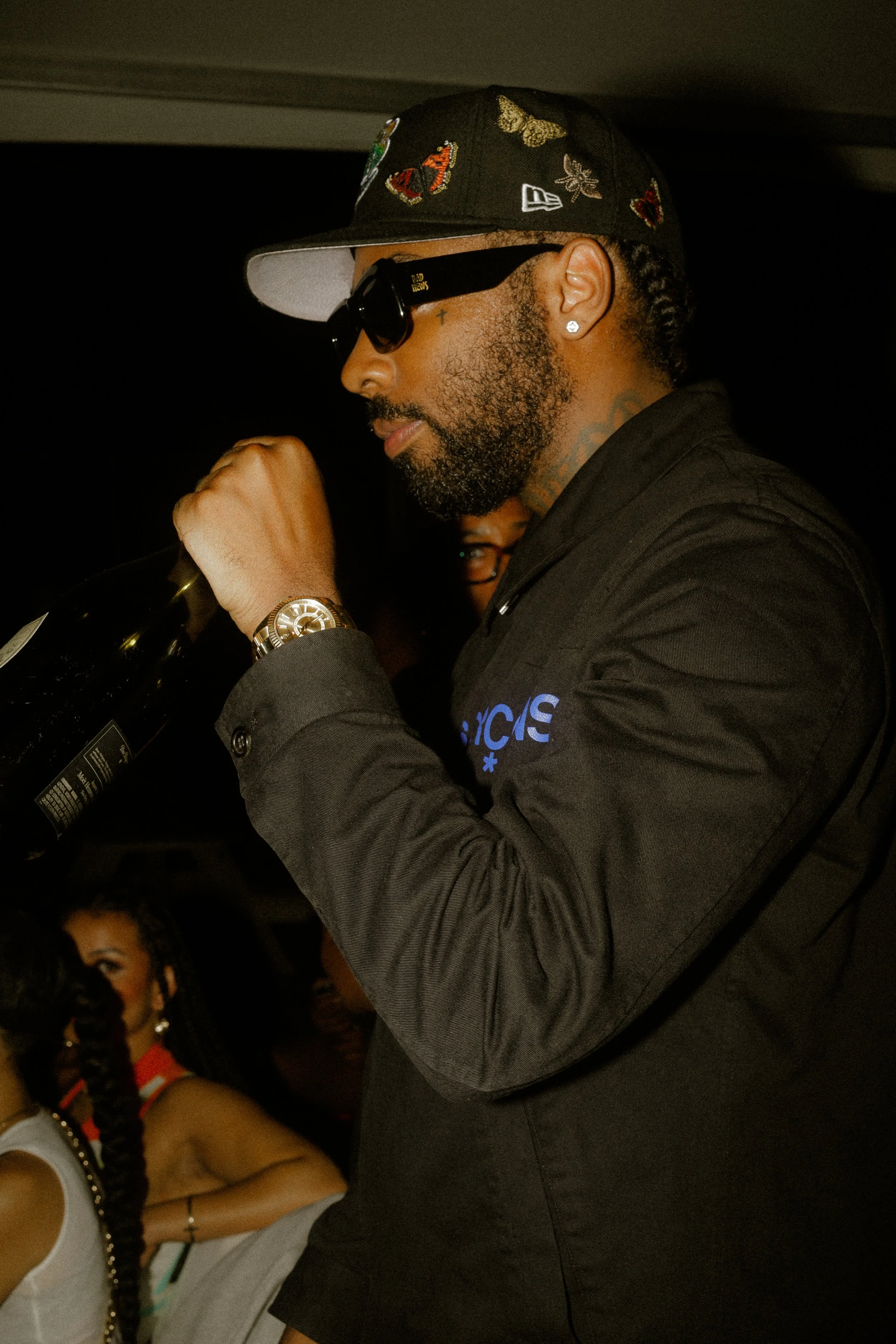 Man in black outfit, cap, and sunglasses holding a bottle at a party.