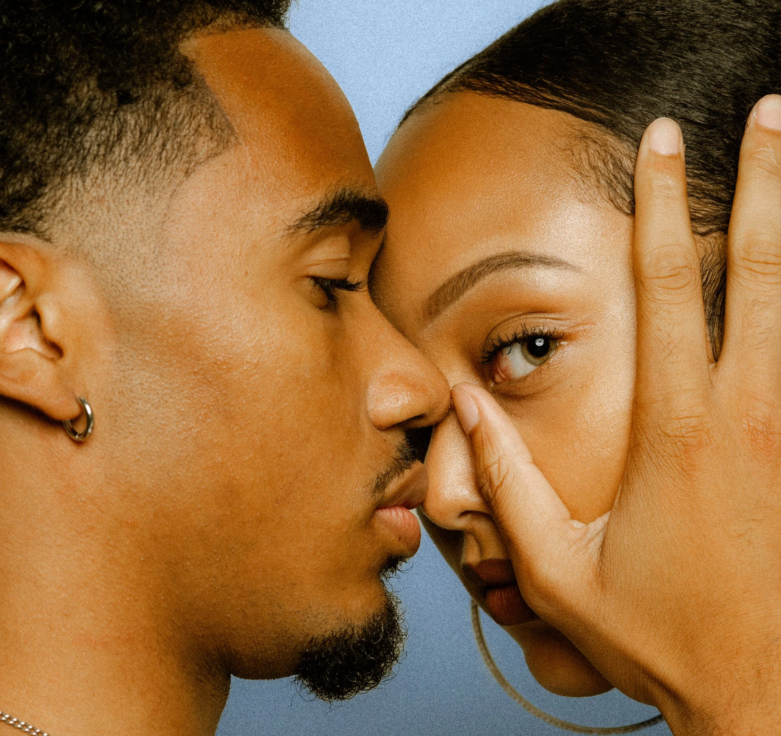 Close-up of two people facing each other with their noses touching. One person's hand frames their eye.