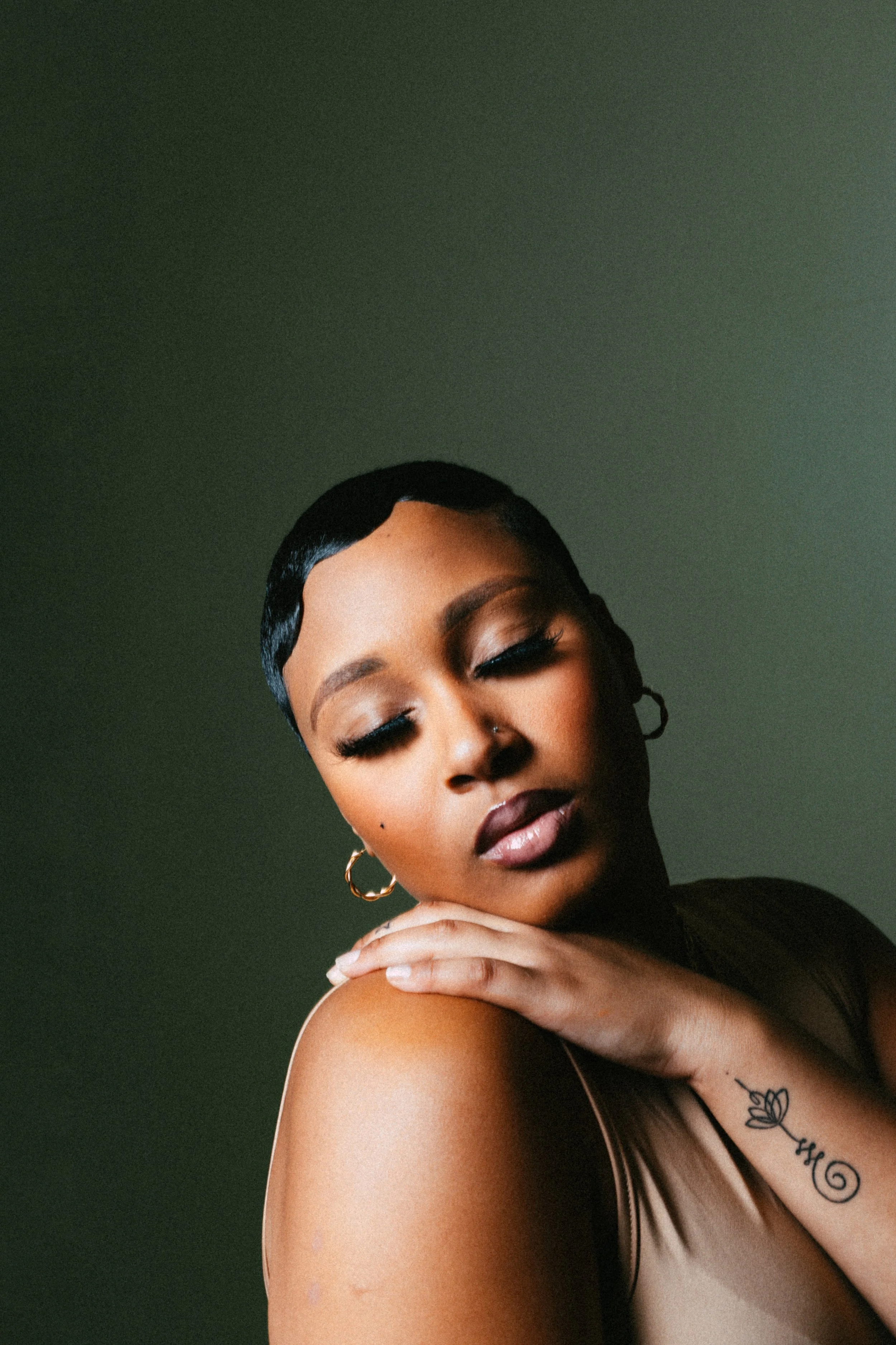 Woman with short hair and hoop earrings, eyes closed, resting hand on shoulder, tattoo visible.