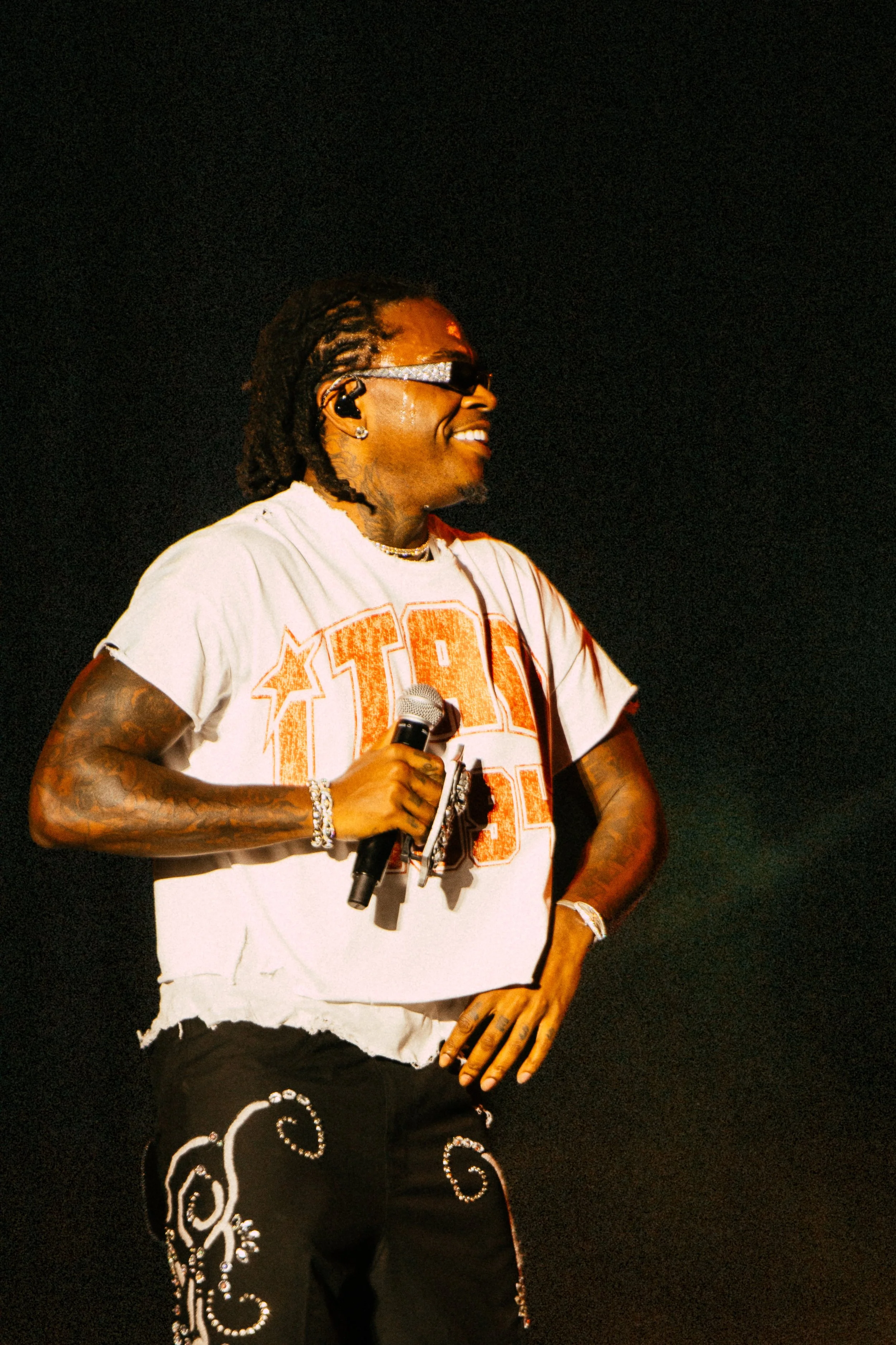 Performer on stage wearing sunglasses and a white graphic t-shirt, holding a microphone with decorative pants and jewelry, smiling under stage lights.