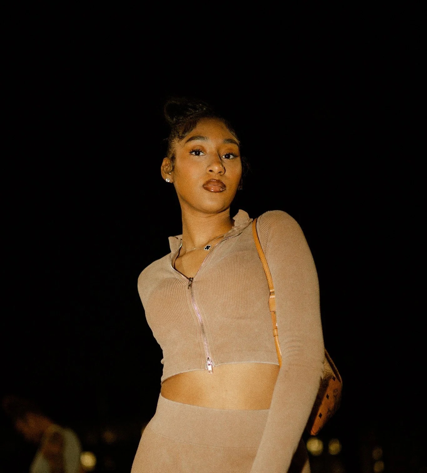 Person wearing a beige zip-up top and matching pants standing against a dark background, carrying a brown shoulder bag.