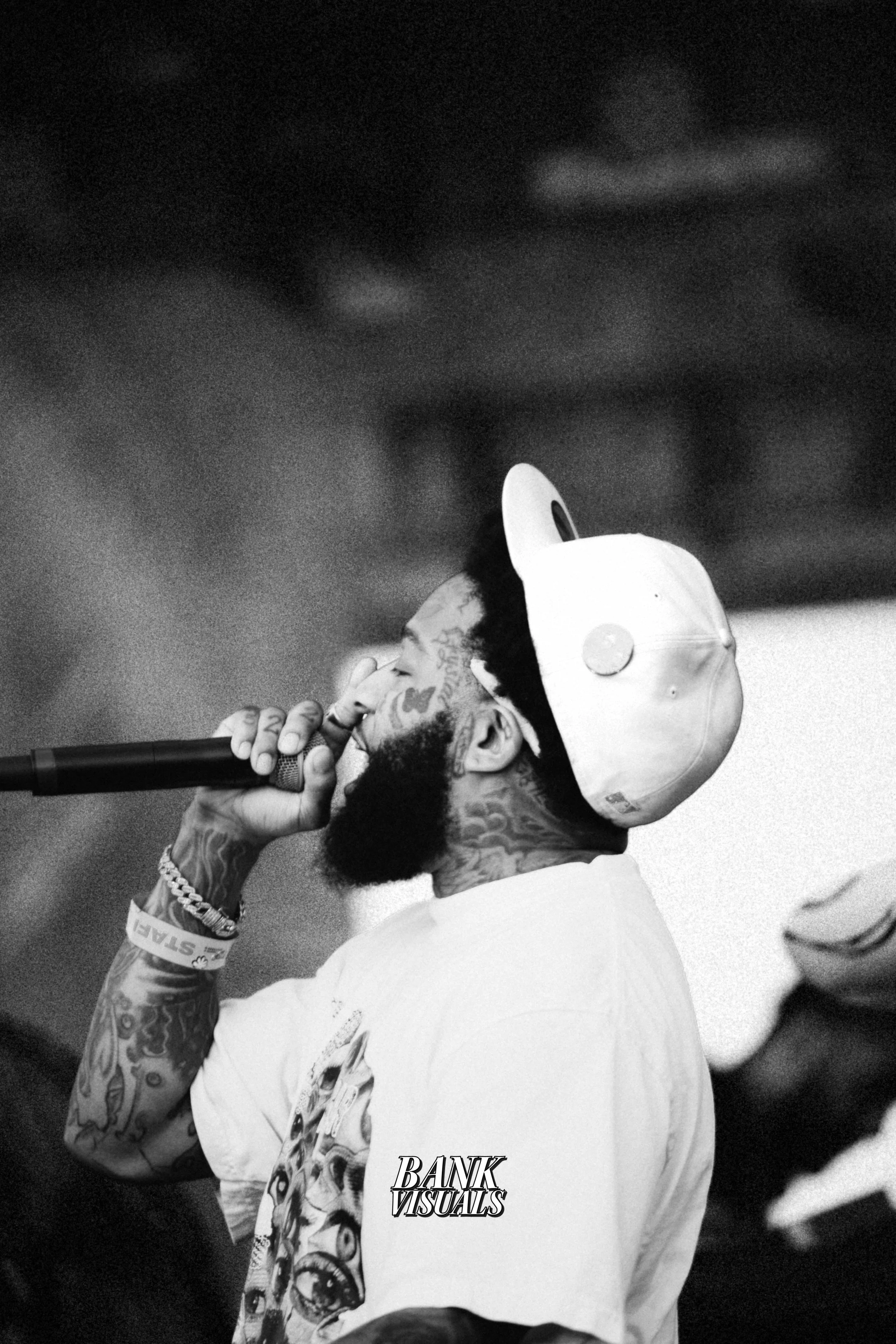 A black and white photo of a tattooed man with a beard, wearing a backward cap and a graphic T-shirt, singing into a microphone with visible intensity.