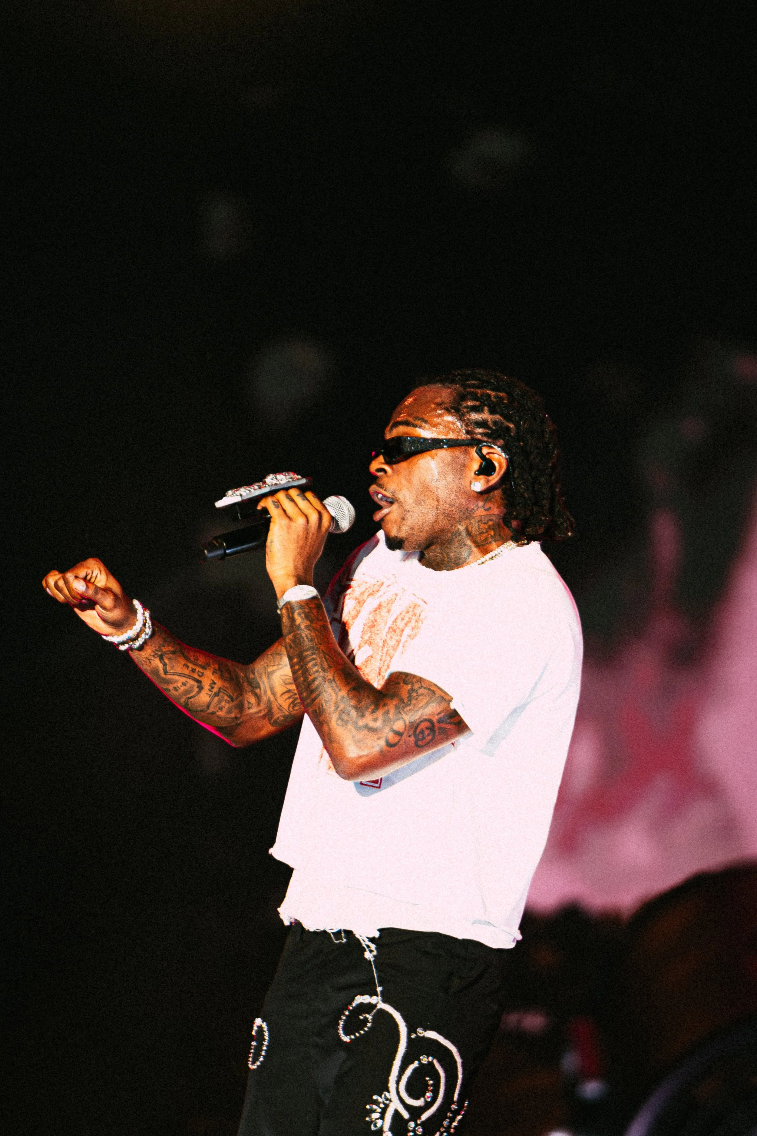Performer on stage holding microphone, wearing sunglasses, white shirt, and patterned black pants.