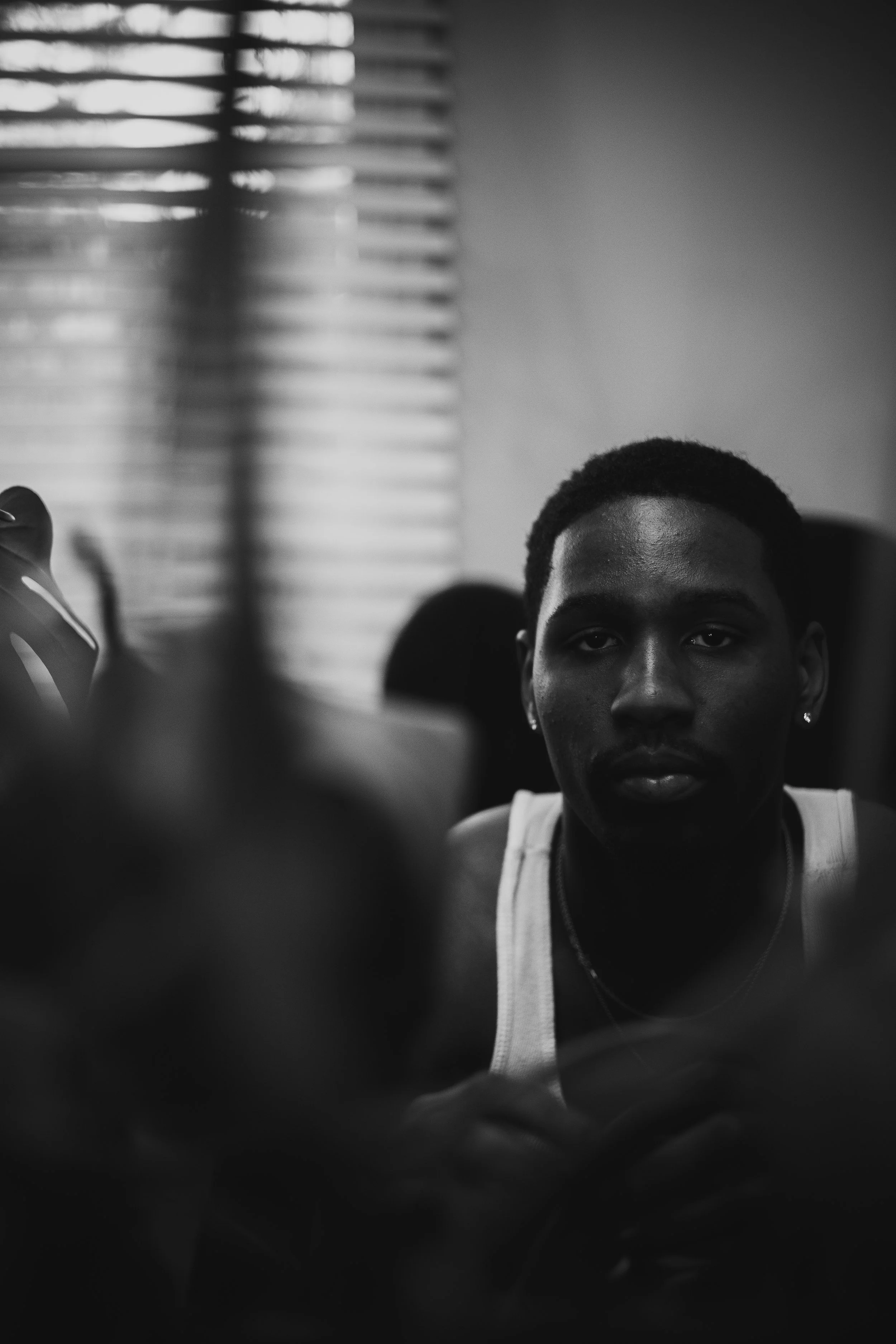 Black and white portrait of a man in a tank top sitting indoors with window blinds in the background.