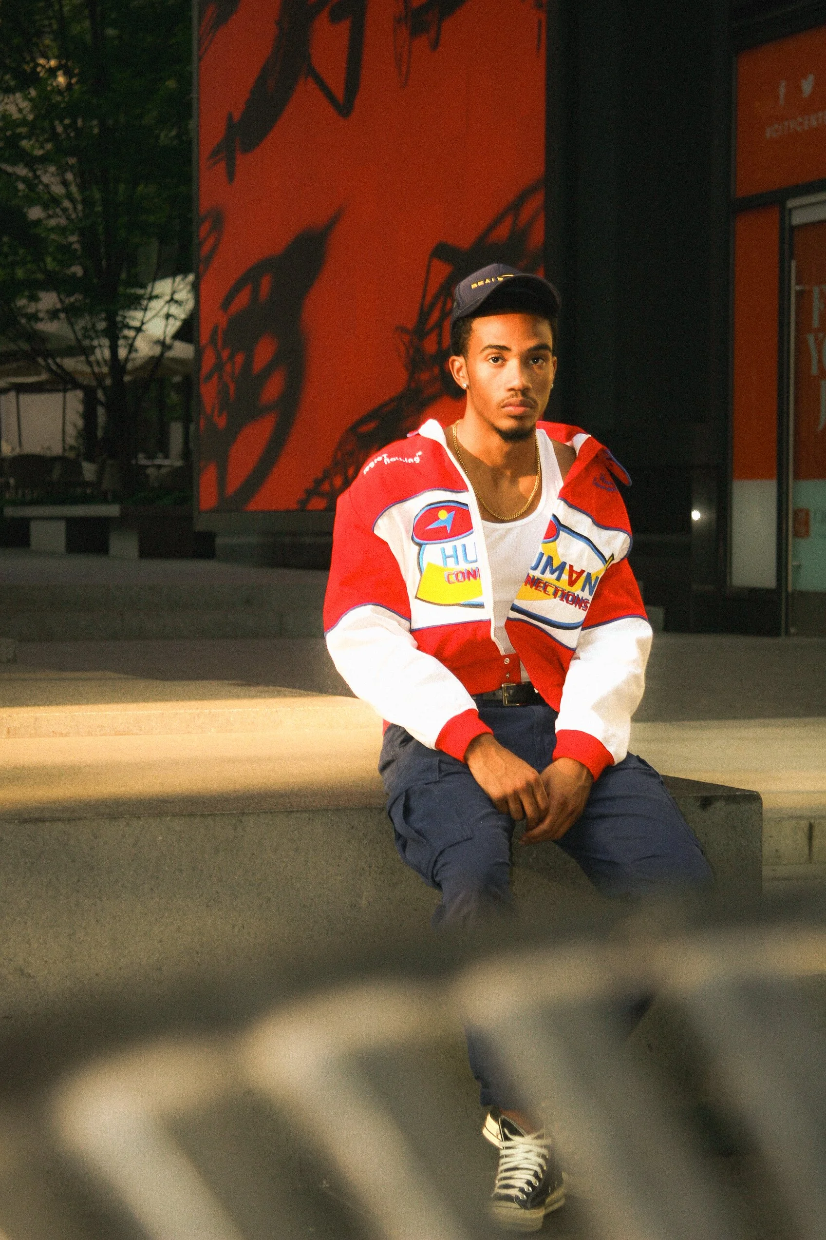 Man sitting outdoors wearing a red and white jacket, blue pants, and a cap.
