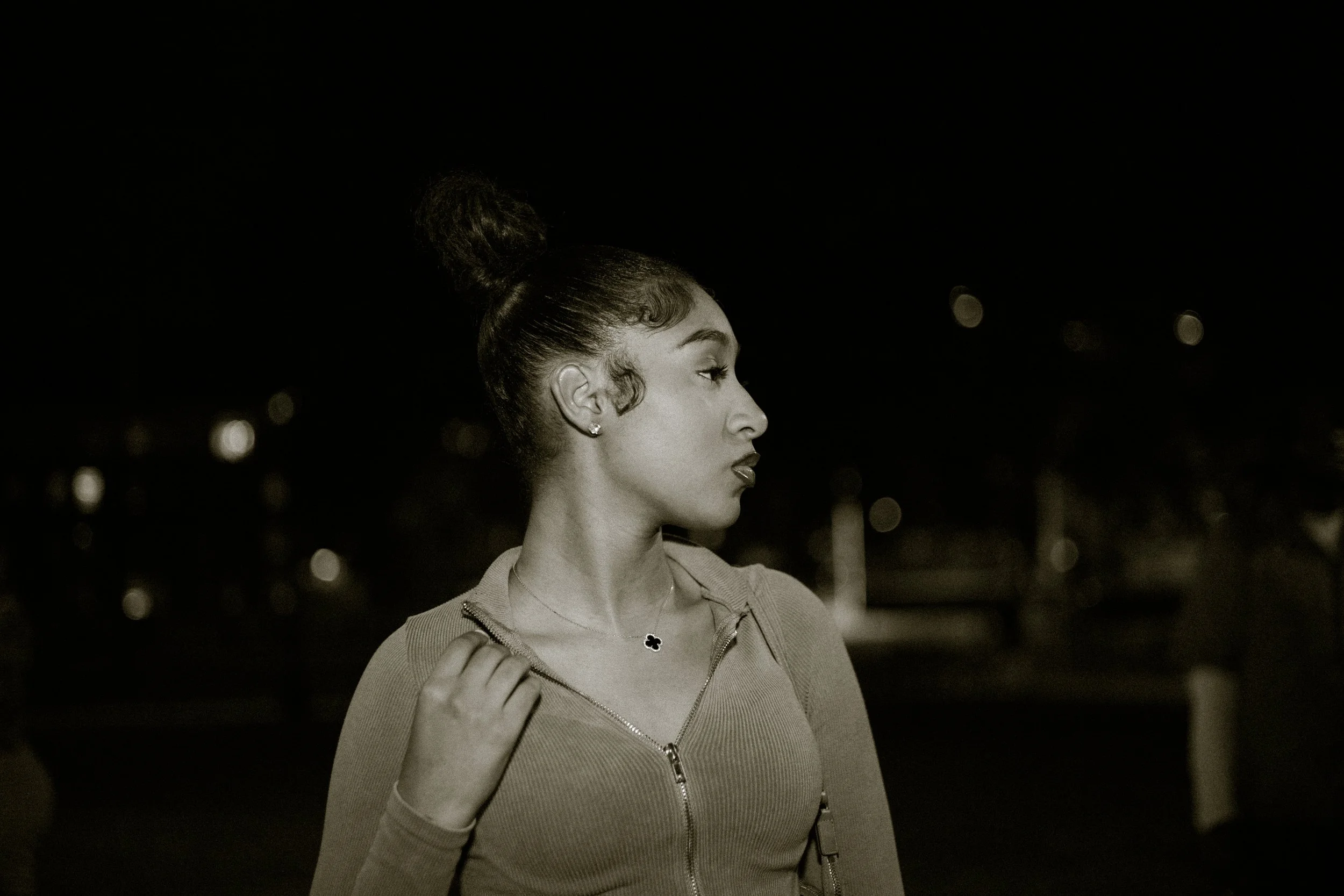 Black and white photo of a woman in profile, wearing a zip-up top and a necklace, set against a dark background with bokeh lights.