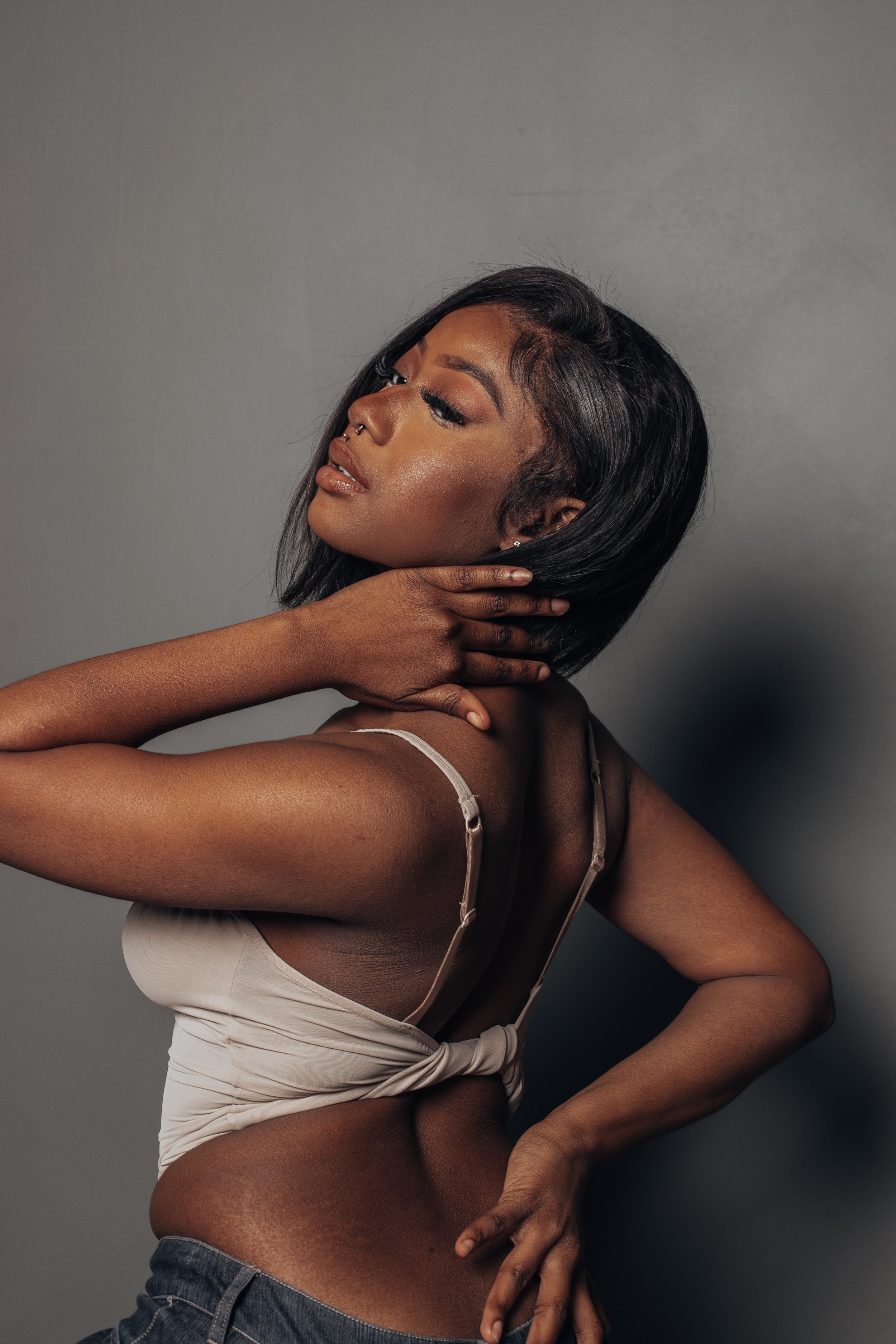 Woman posing with hand on her neck, wearing a beige top and dark pants, against a gray background.