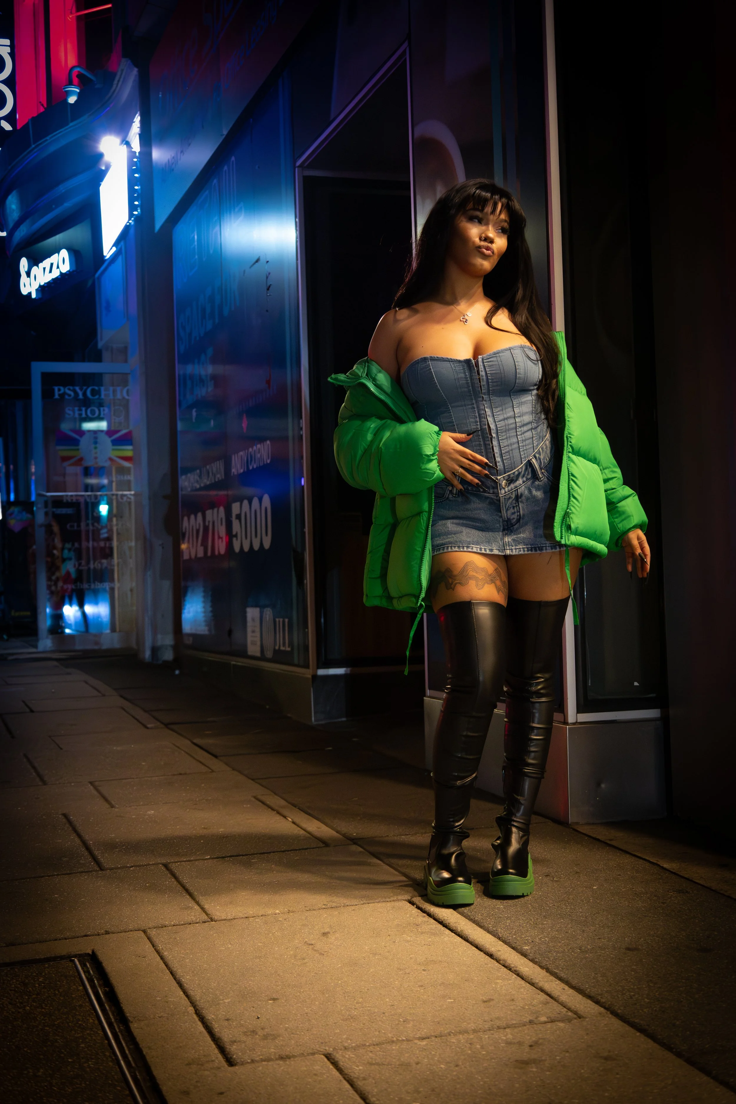 A woman in a green jacket and denim outfit walking on a city street at night, wearing black thigh-high boots.