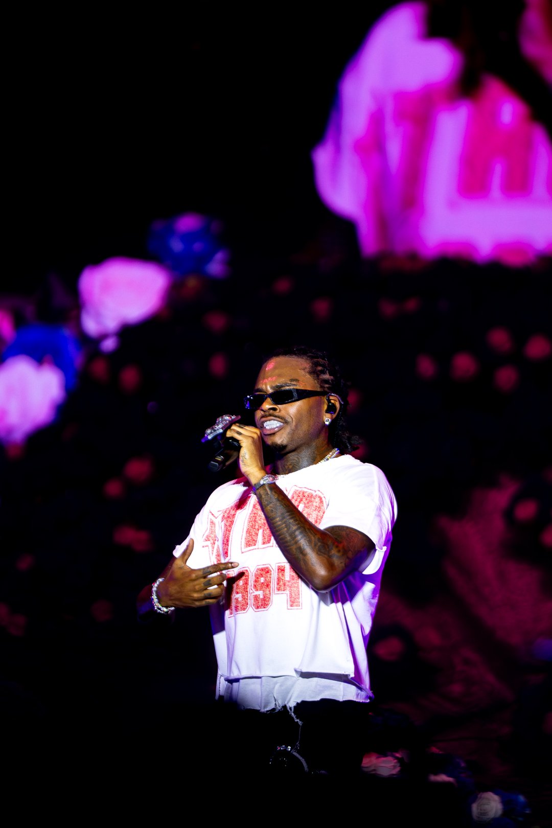 Performer singing on stage, wearing sunglasses and a white t-shirt, holding a microphone.