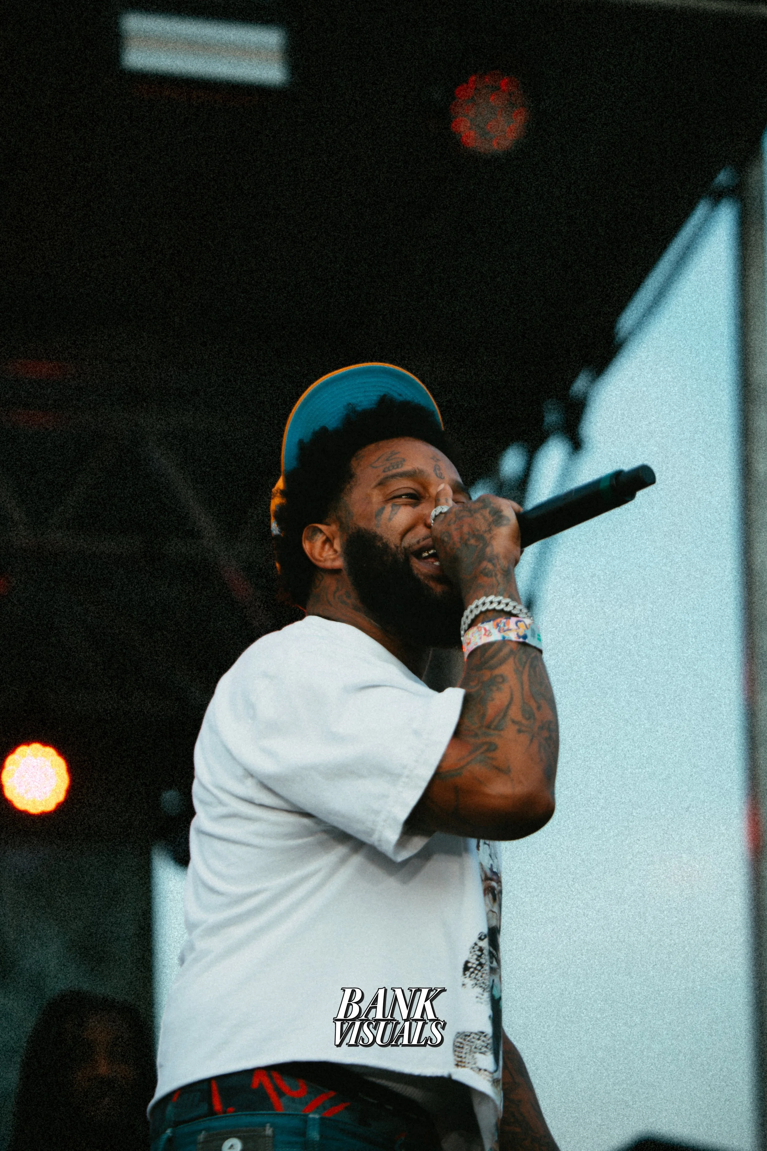 A man with tattoos performs on stage, wearing a white t-shirt and a blue cap, holding a microphone. The background features stage lighting.