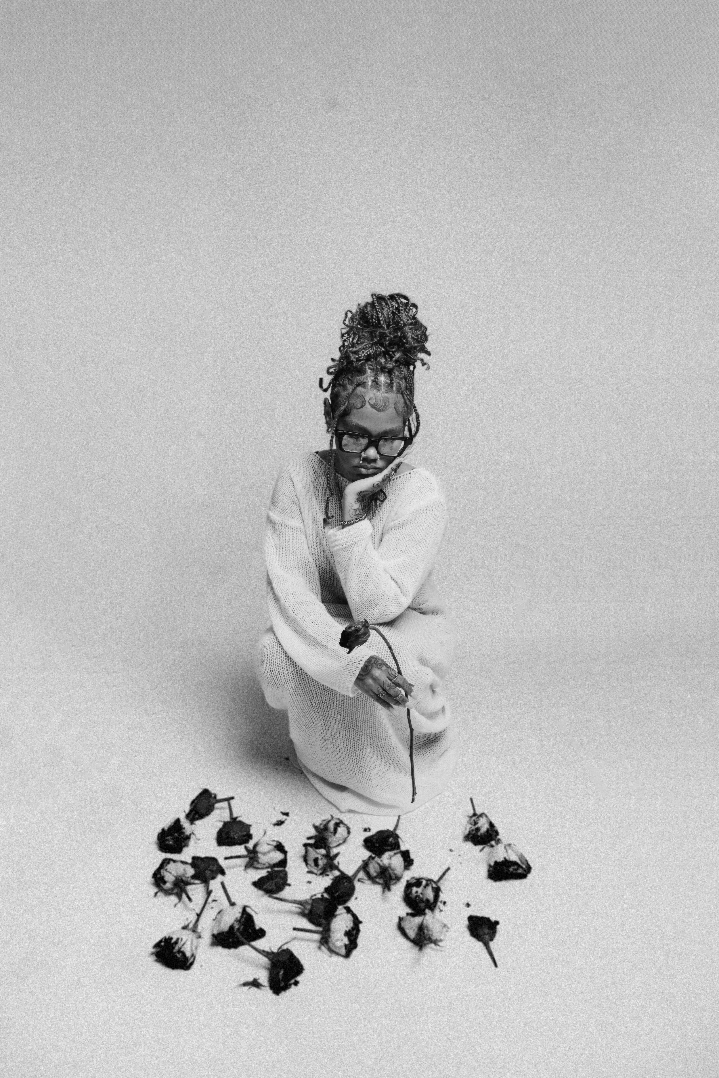 Black and white photo of a person in a sweater with braided hair sitting and holding a single flower, surrounded by scattered withered roses.
