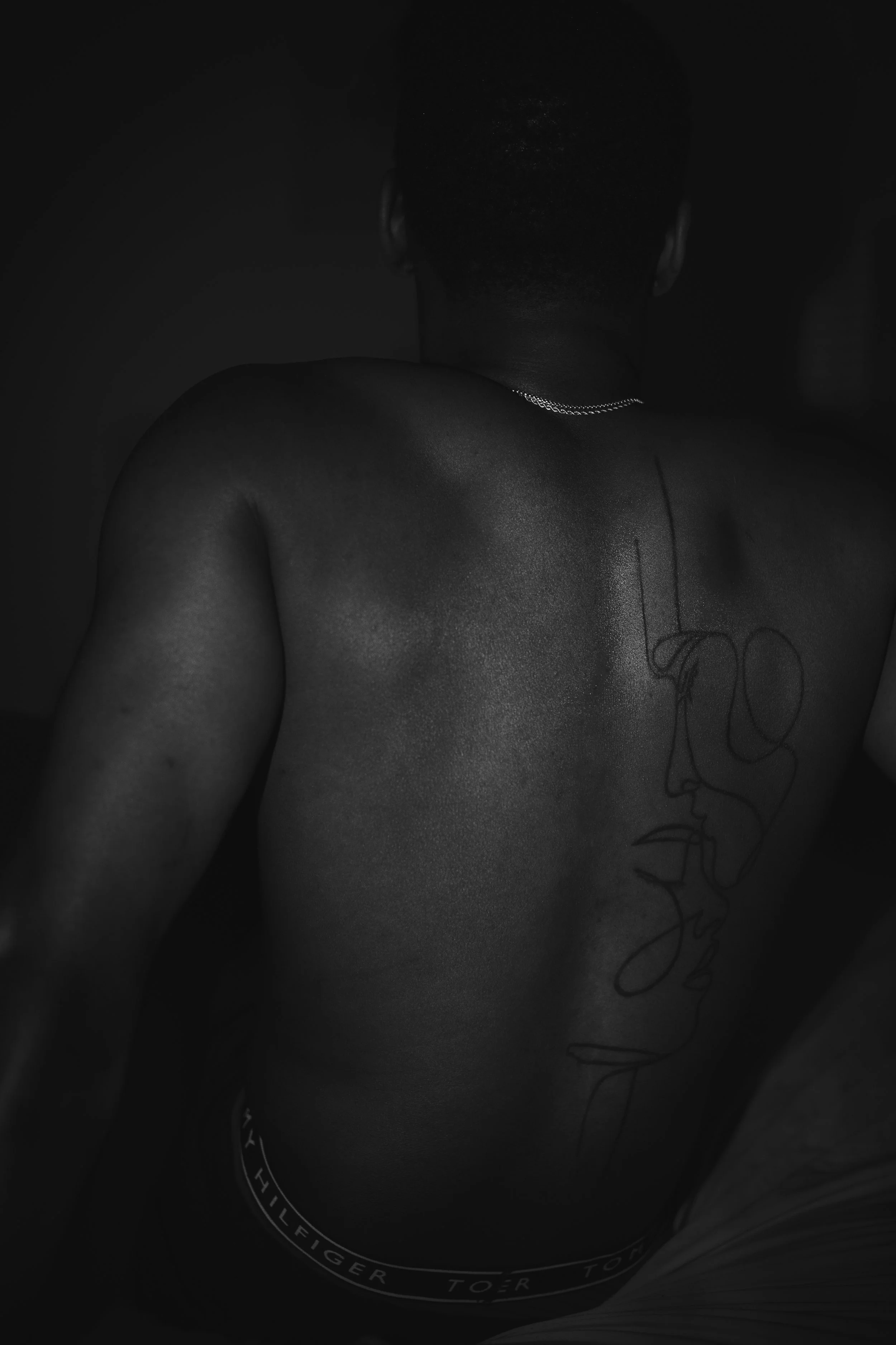 Black and white photo of a person's back with a line-art tattoo of a face, wearing a necklace and dark underwear.