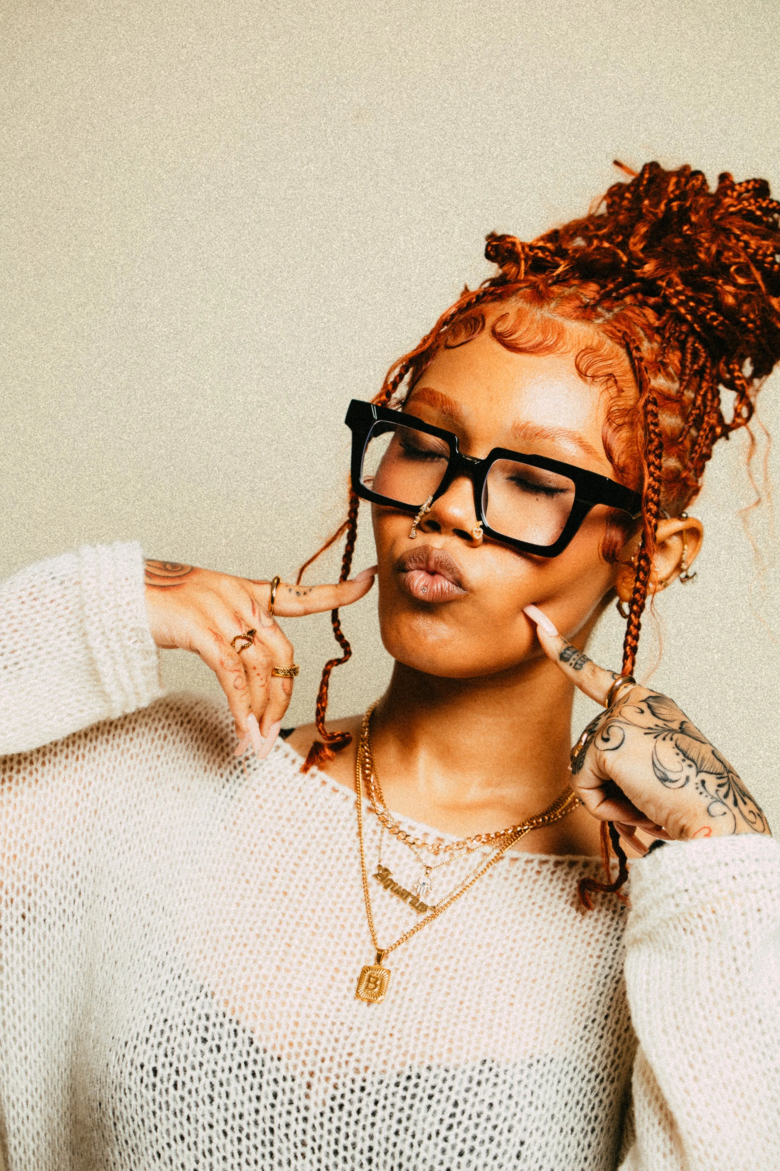 Person with braided hair, wearing glasses, multiple necklaces, and tattoos on their hands, posing with fingers on cheeks.