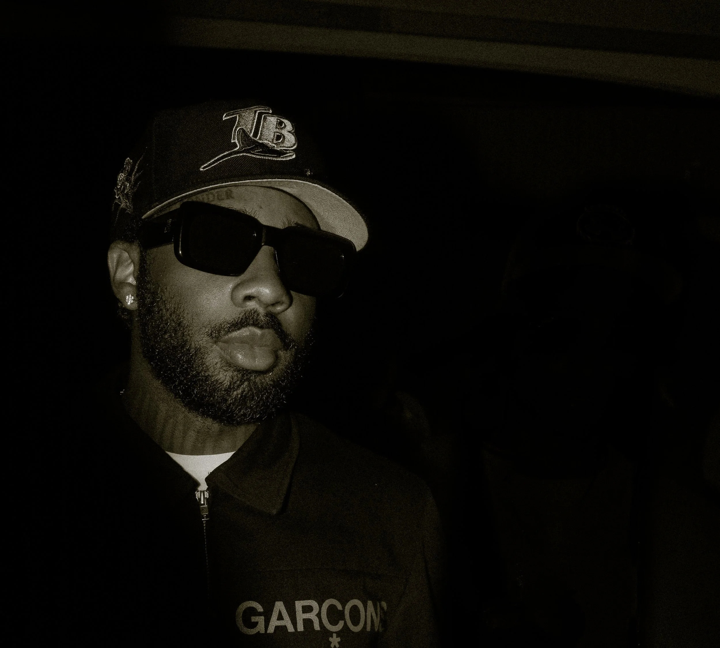 Person wearing a baseball cap and sunglasses in a dark setting.