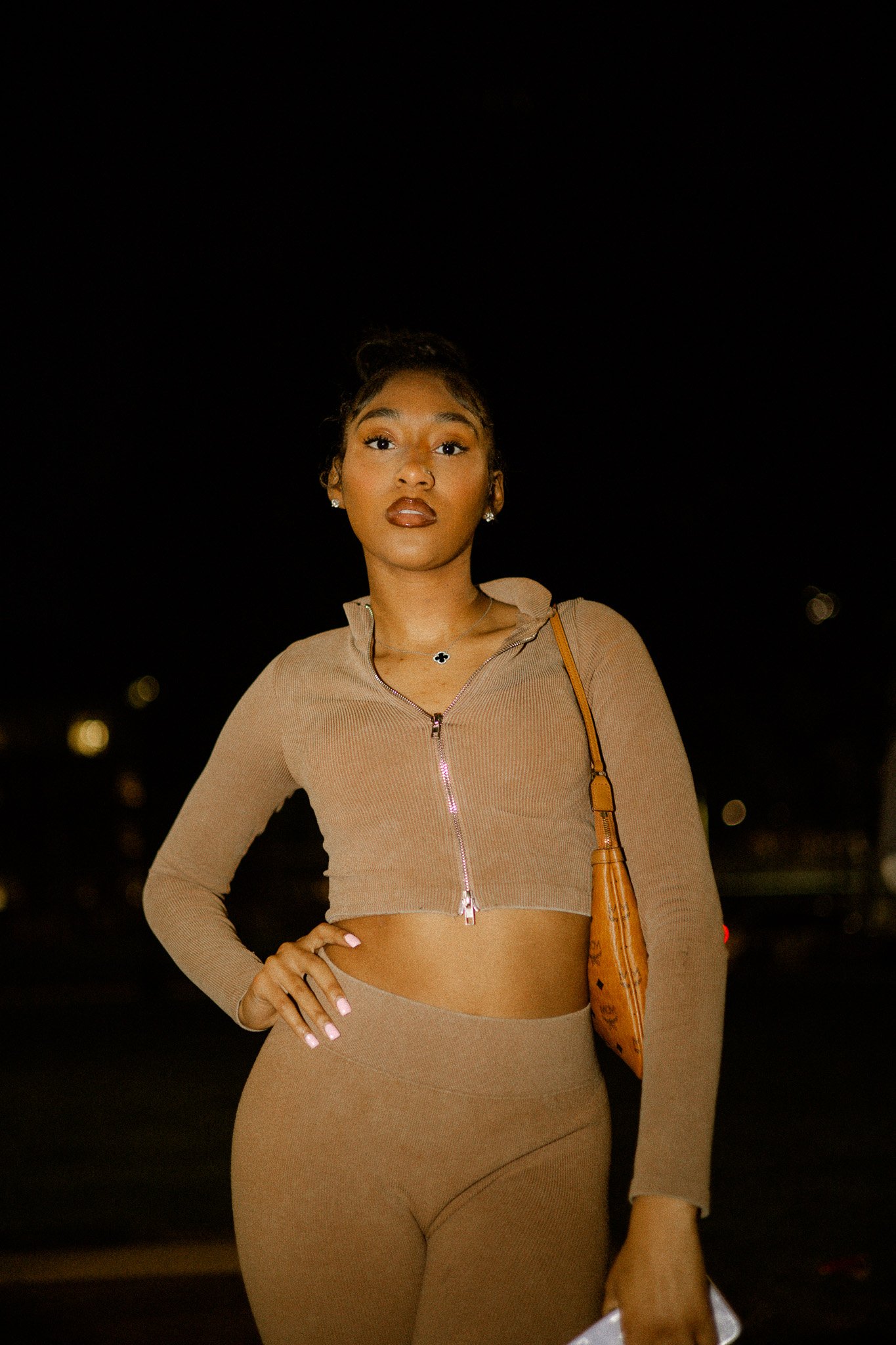 Woman wearing a brown cropped jacket and pants, holding handbag, standing at night.