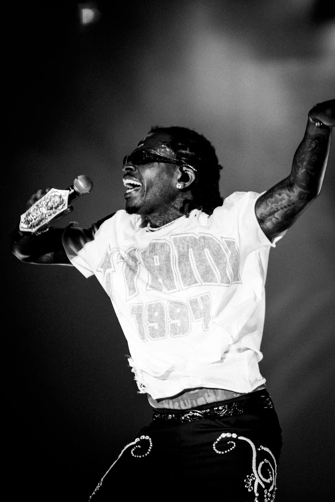 Black and white photo of a person singing passionately on stage, holding a microphone decorated with diamonds, wearing sunglasses and a shirt with '1994' printed on it.