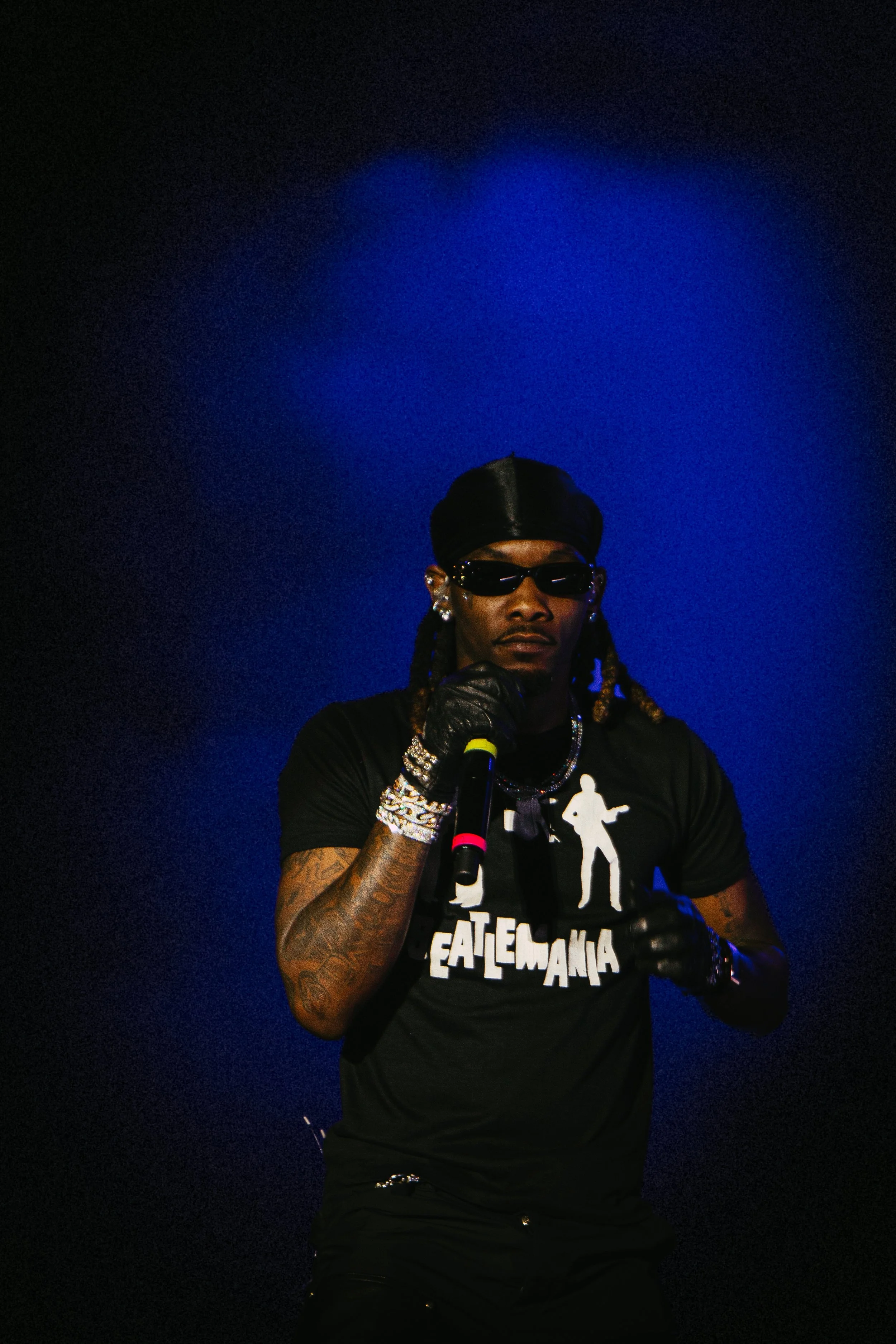 Performer on stage wearing sunglasses and a black shirt with "EATEMANIA" design, holding a microphone, in front of a blue background.