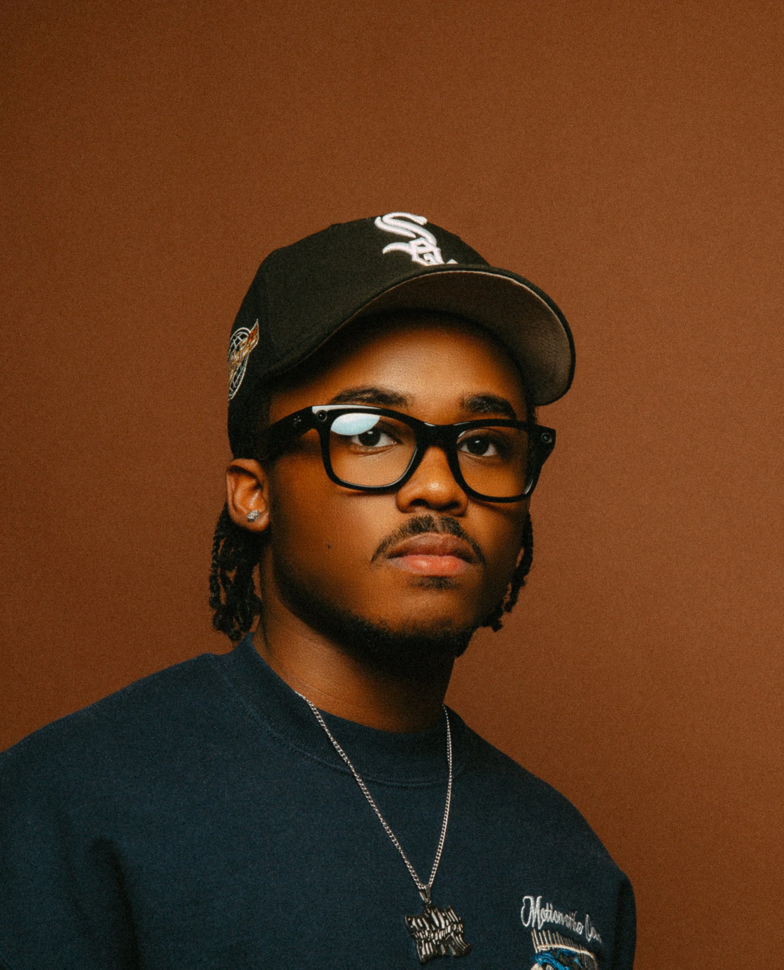 Person wearing glasses and a baseball cap against a brown background.