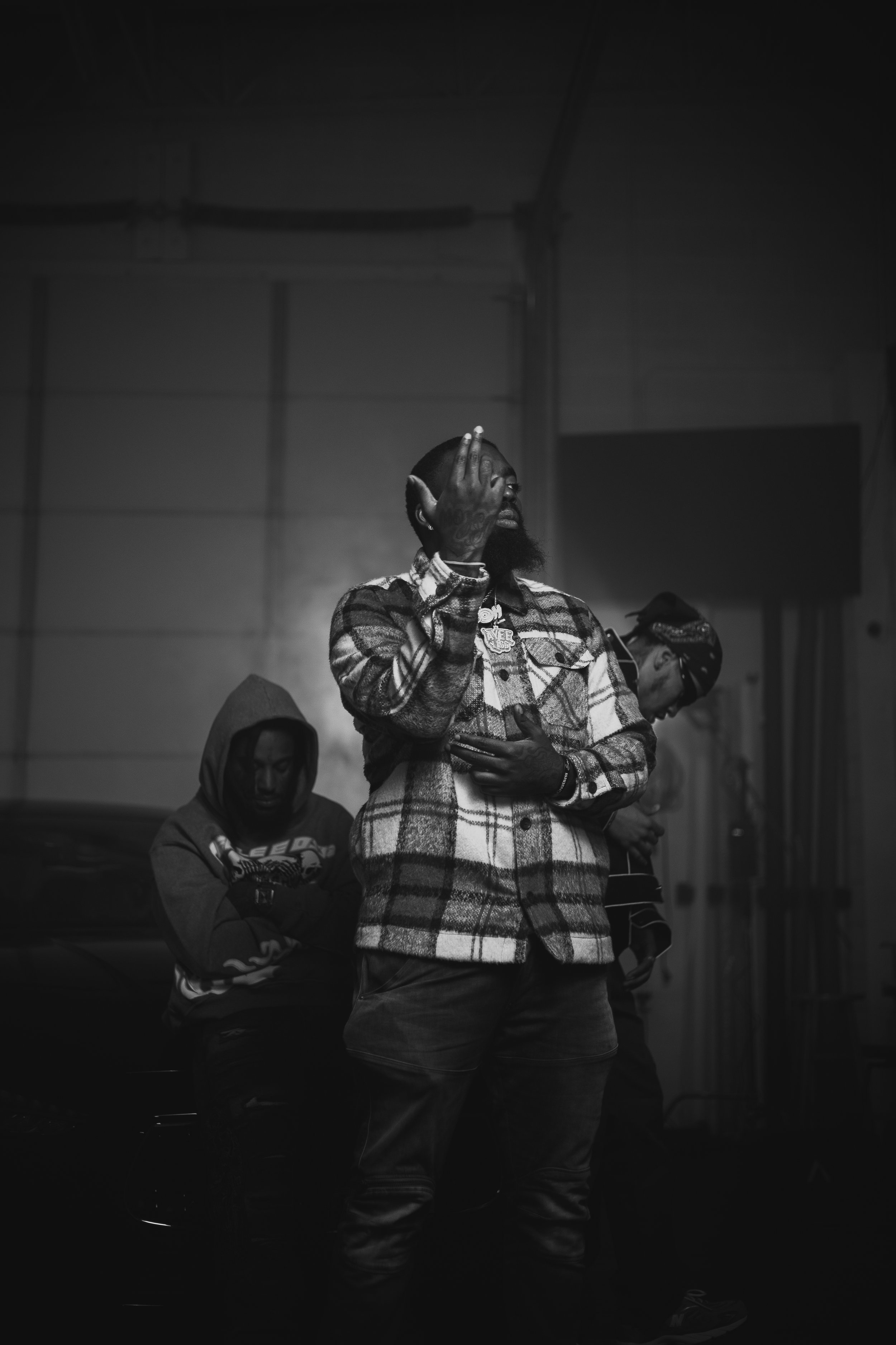 Black and white photo of three individuals; one in the foreground wearing a plaid jacket, gesturing with their hand, and two in the background, one wearing a hoodie and another a bandana.