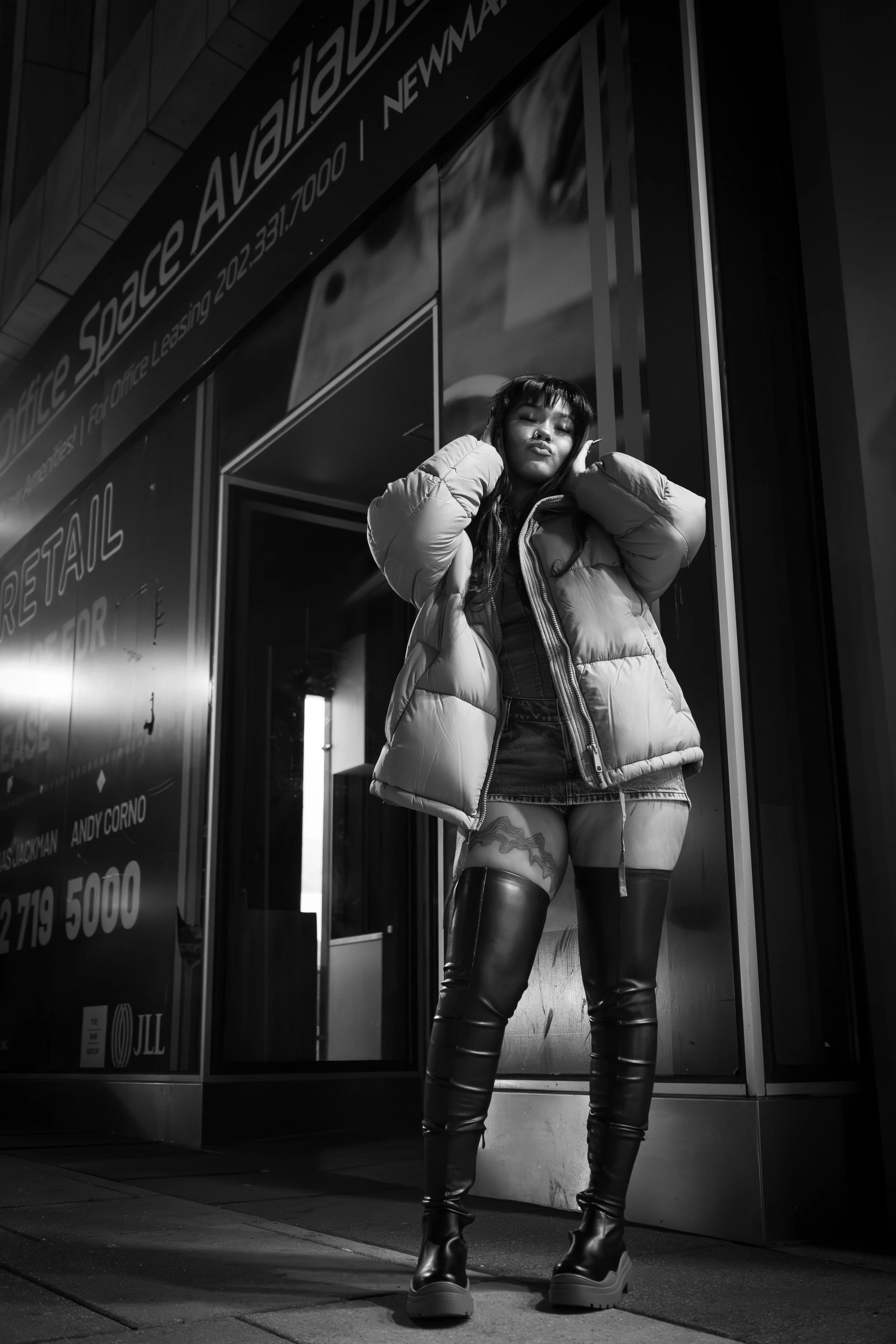 Black and white photo of a woman in a puffy jacket and thigh-high boots standing outside a retail space with "Office Space Available" signage in the background.