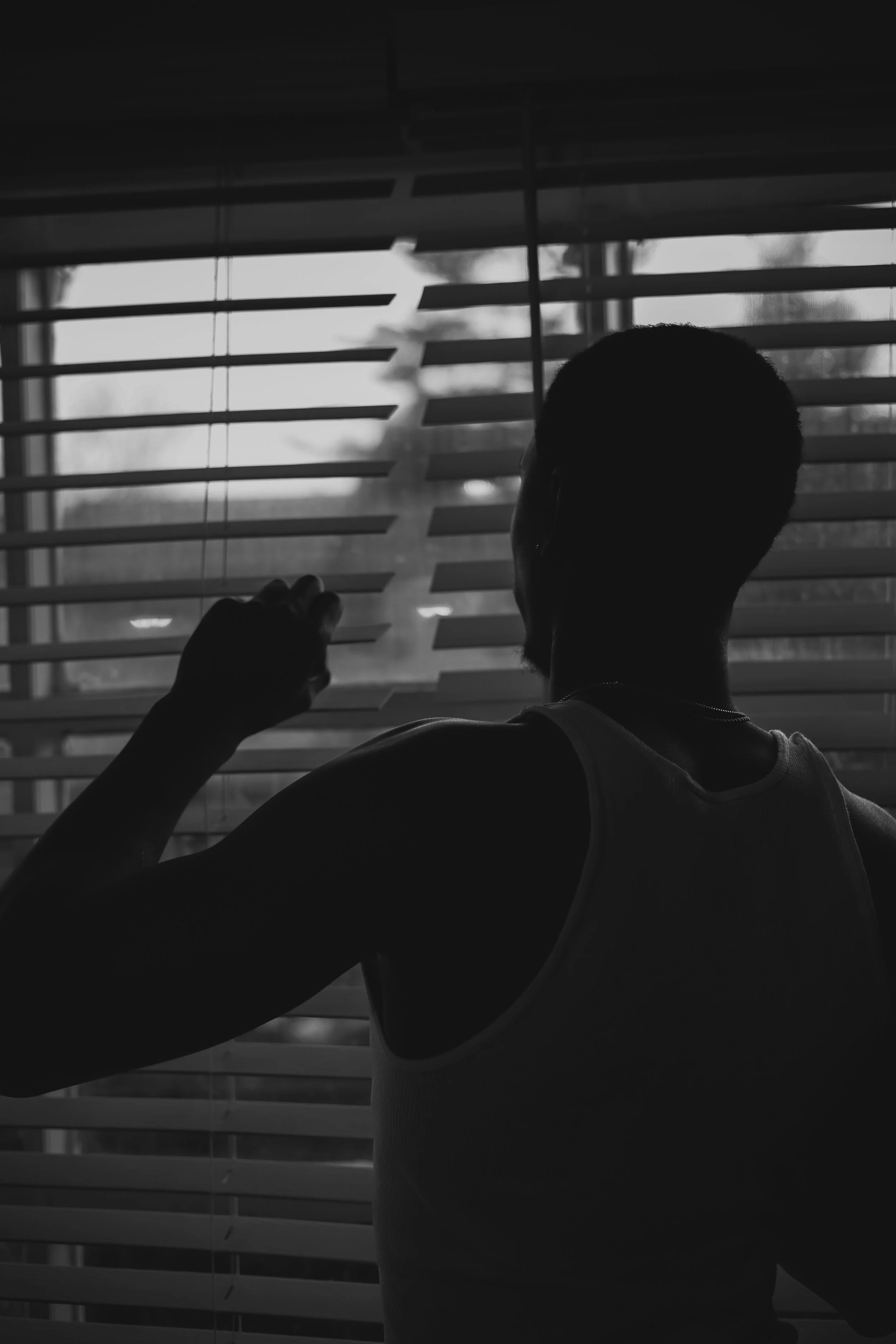Silhouette of a person in a tank top peering through window blinds.