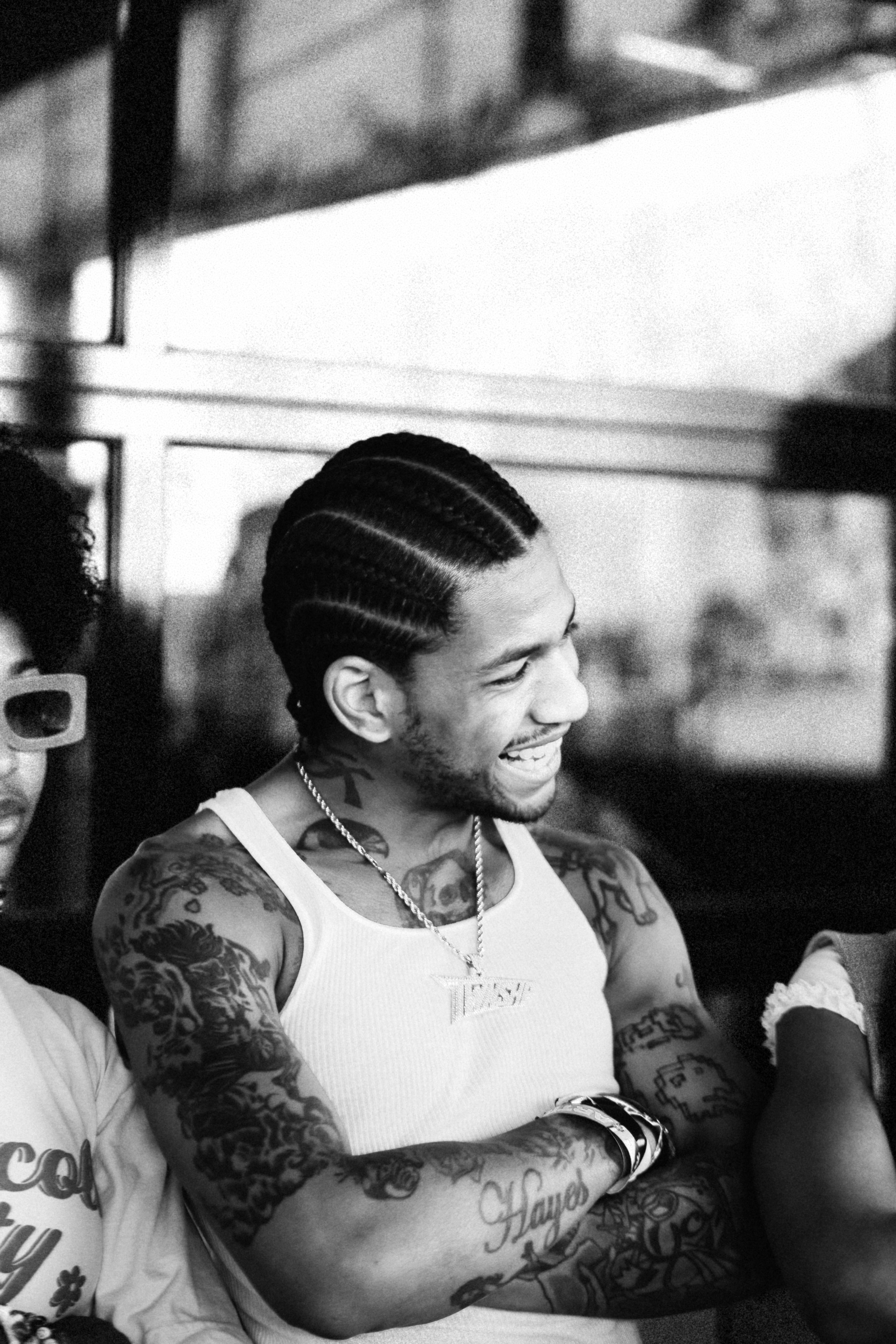 Black and white photo of a smiling person with braided hair, wearing a tank top and necklaces, with visible tattoos on their arms and chest.
