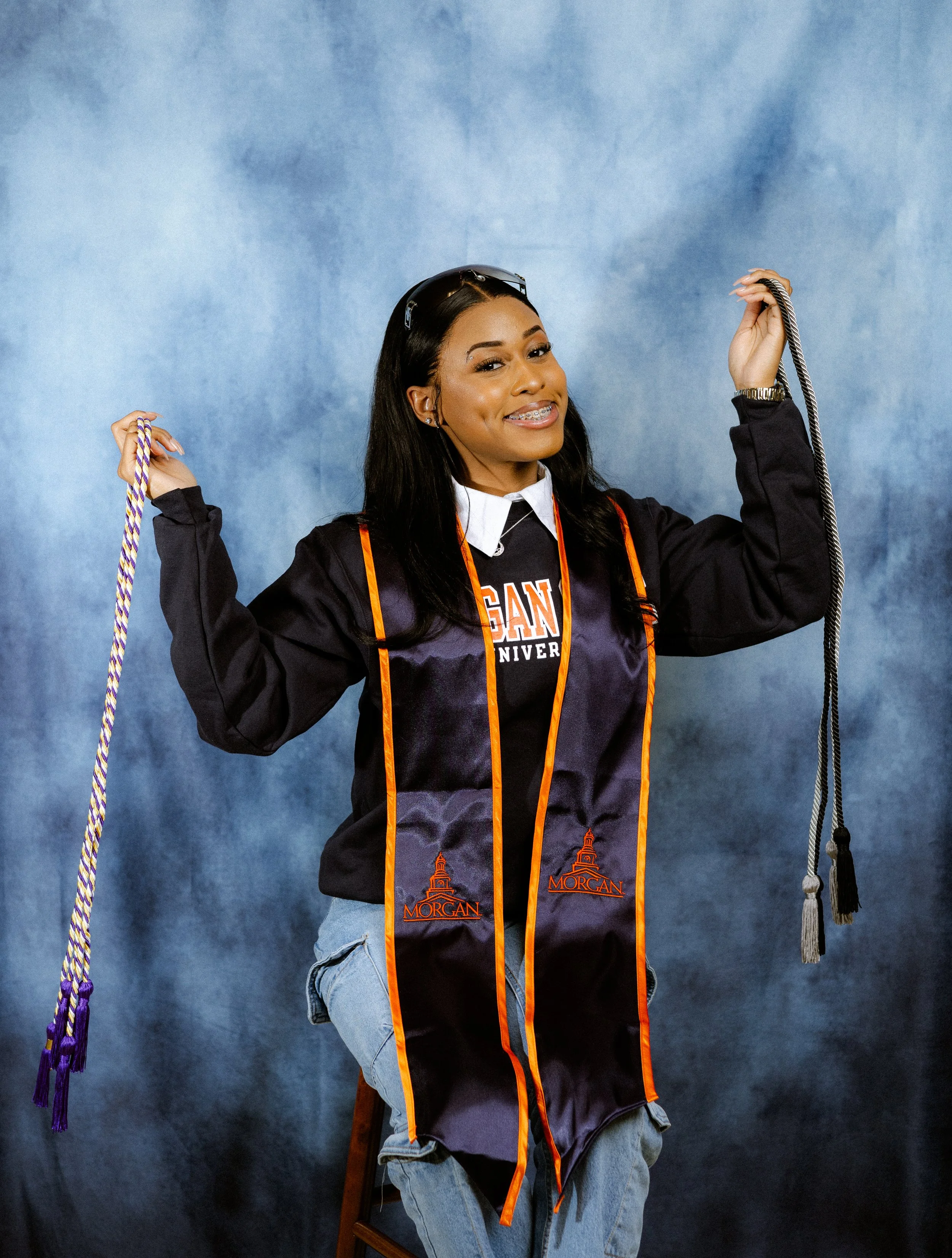 Graduate wearing a Morgan University stole holding honor cords against a blue background.