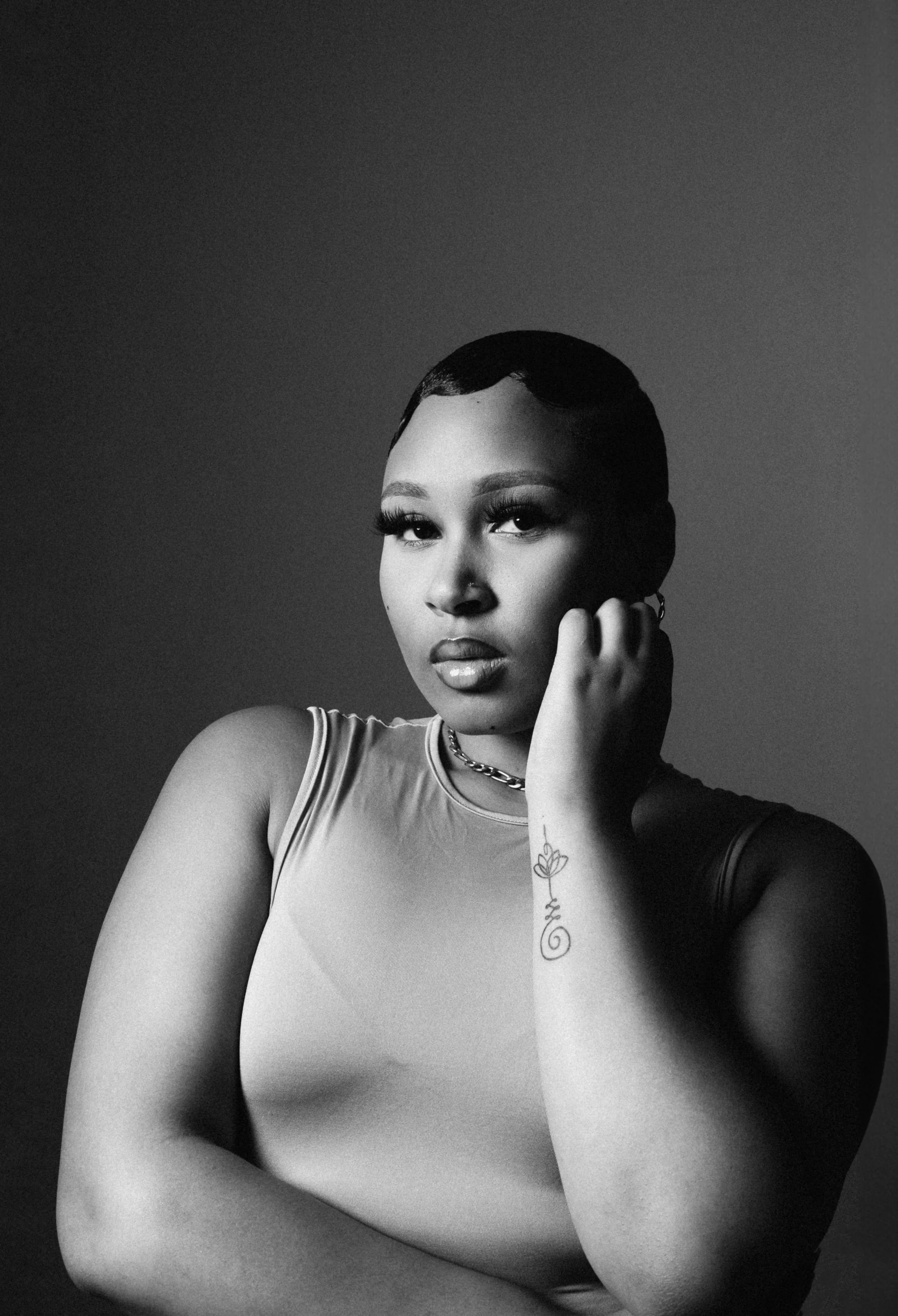 Black and white portrait of a person with short hair, wearing a sleeveless top and a chain necklace, posing with a hand near their face. Visible arm tattoo of a flower design.