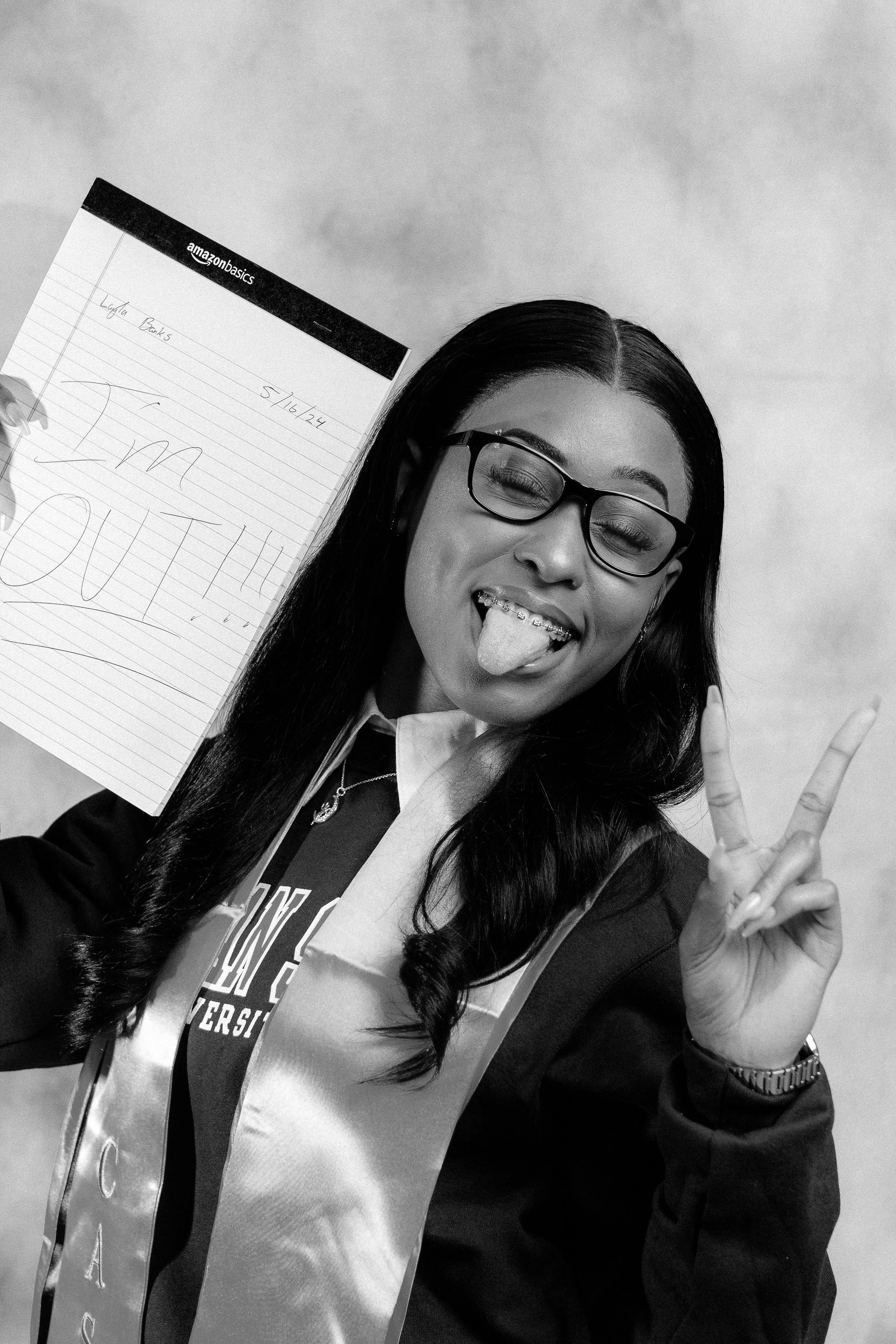 Person wearing glasses and a graduation stole, holding a notepad with "I'm OUT!!!" written on it, showing a peace sign and sticking out tongue.