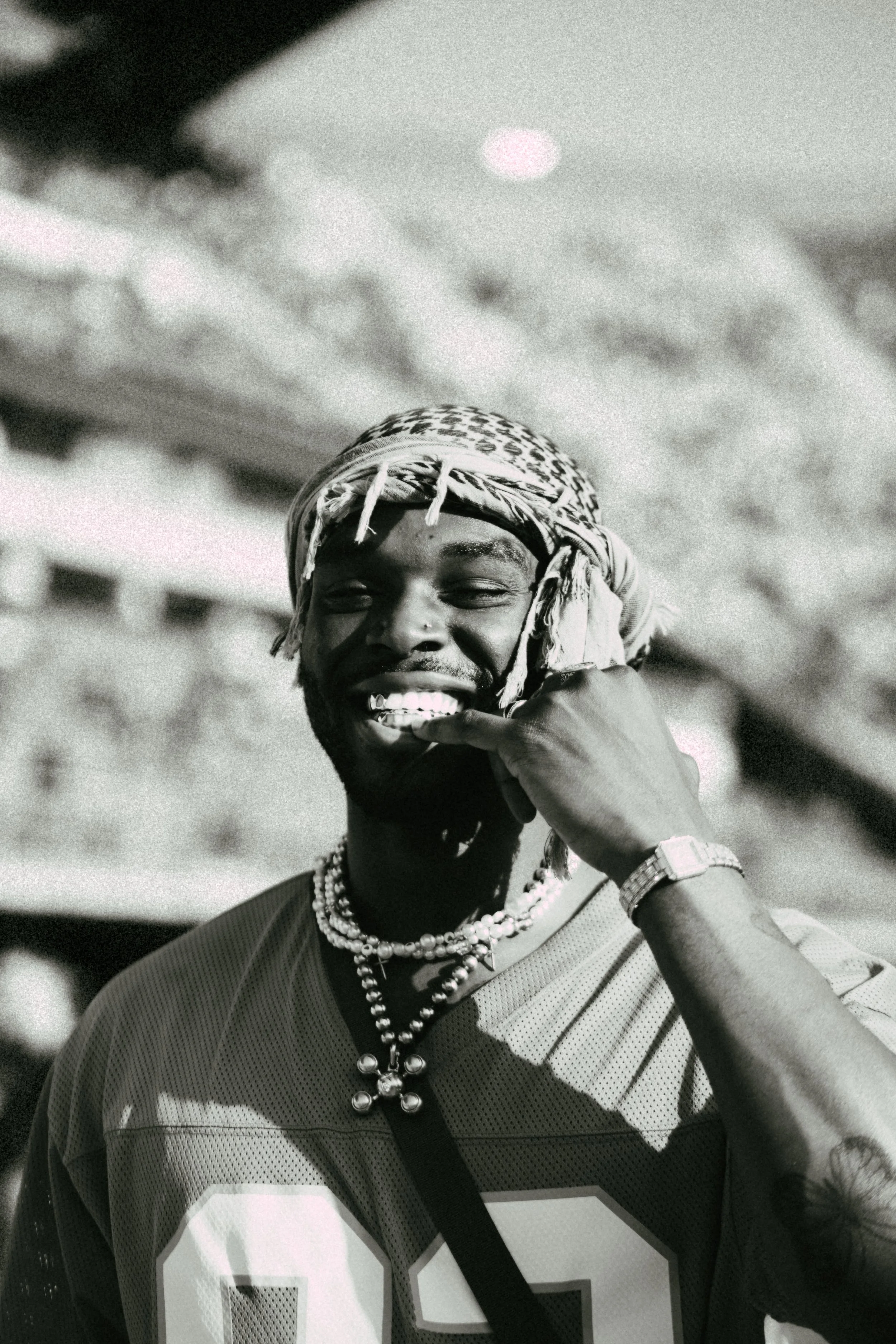 A man smiling, wearing a patterned headscarf, jersey, and jewelry, playfully biting his finger in a stadium setting.