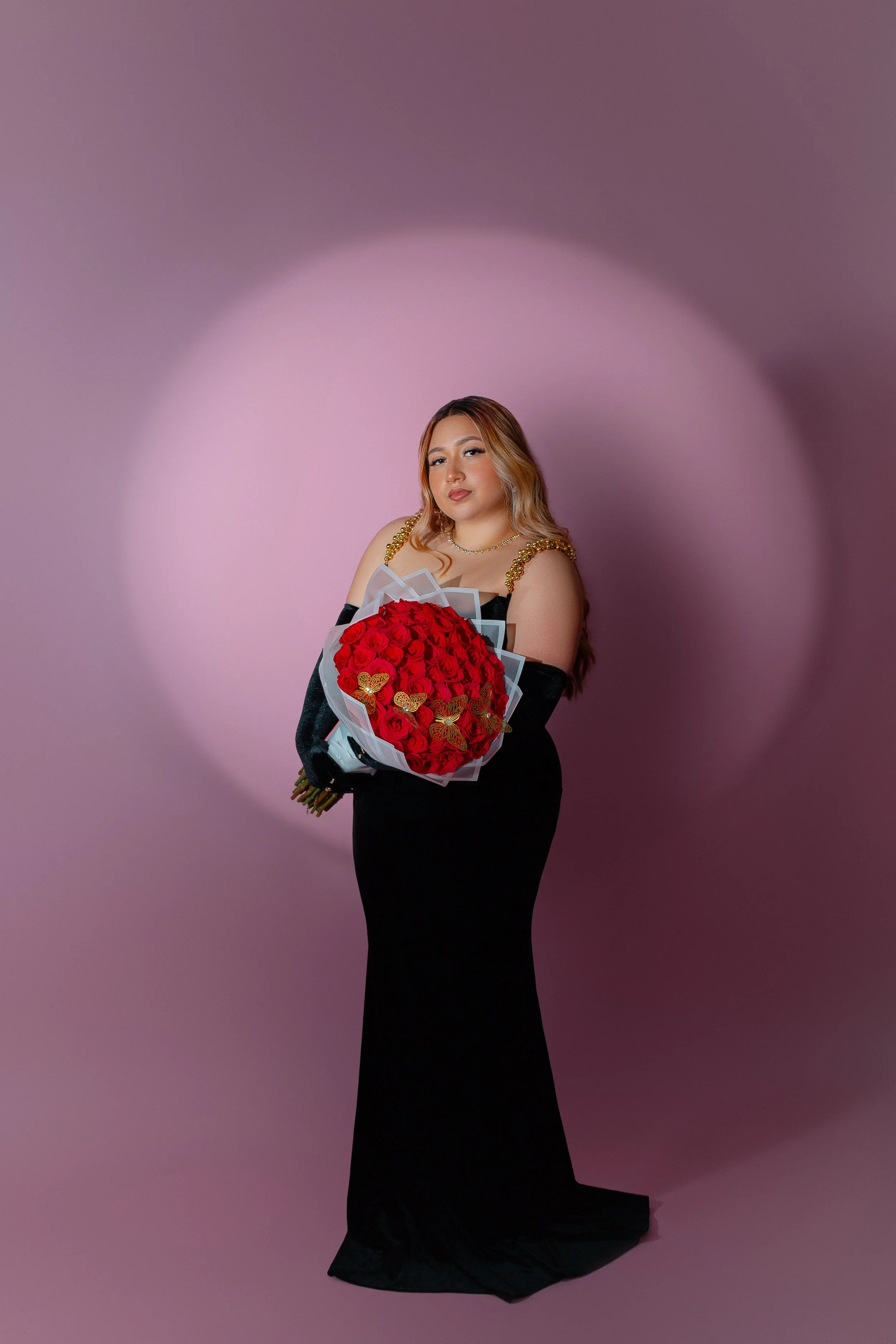 Person in a black dress holding a bouquet of red roses against a pink background.