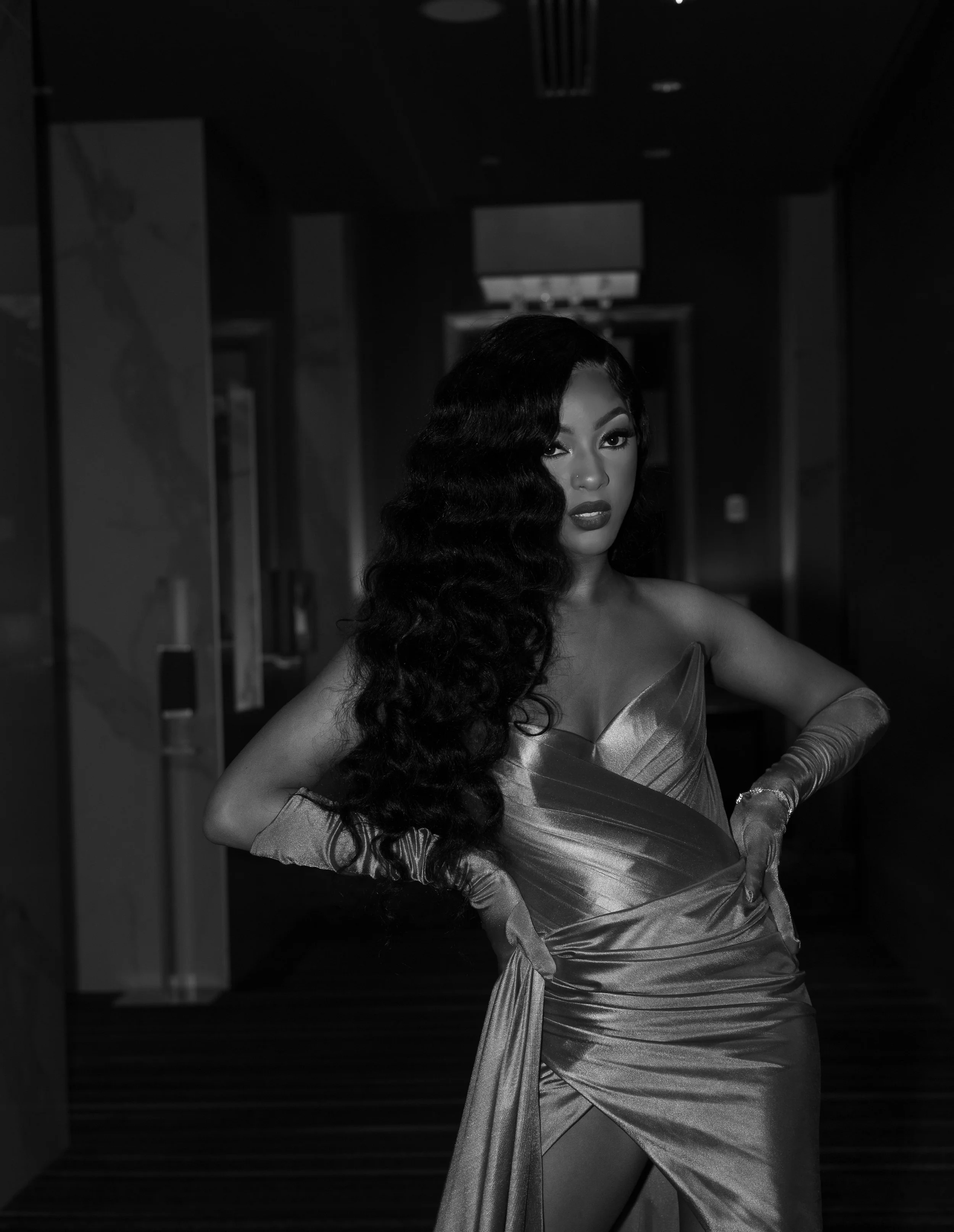 Black and white photo of a person wearing a form-fitting, metallic gown with long gloves, posing confidently in a dimly lit hallway.