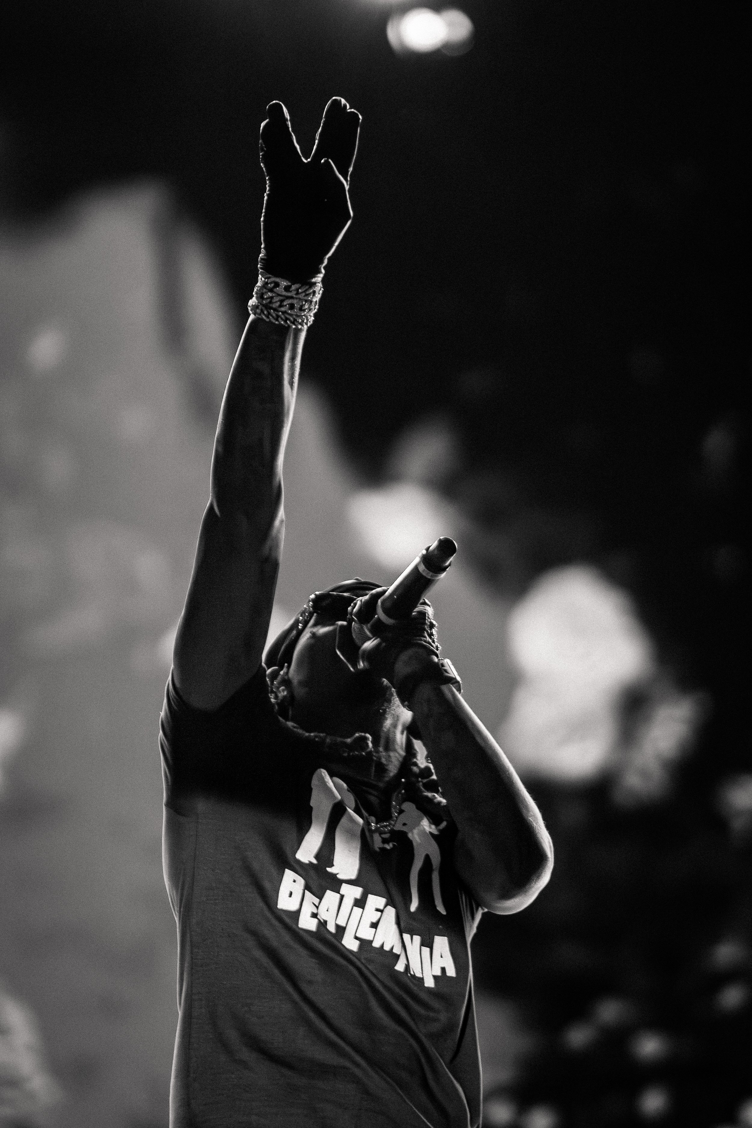 Black and white photo of a performer on stage, wearing a "Beatlemania" T-shirt, holding a microphone and raising one arm with a glove in a gesture.