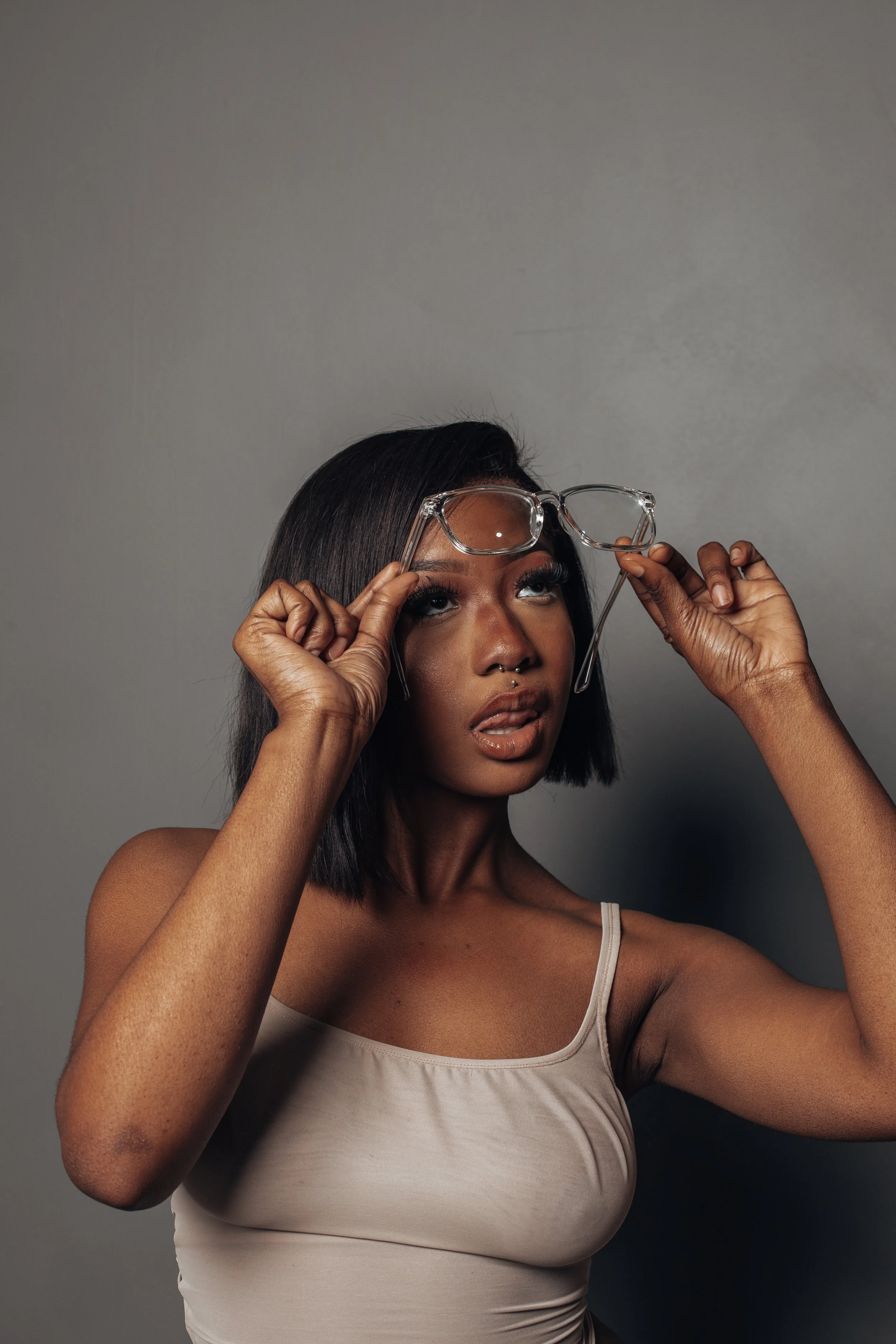 Woman with short hair holding eyeglasses and looking upward, wearing a beige tank top, against a gray background.