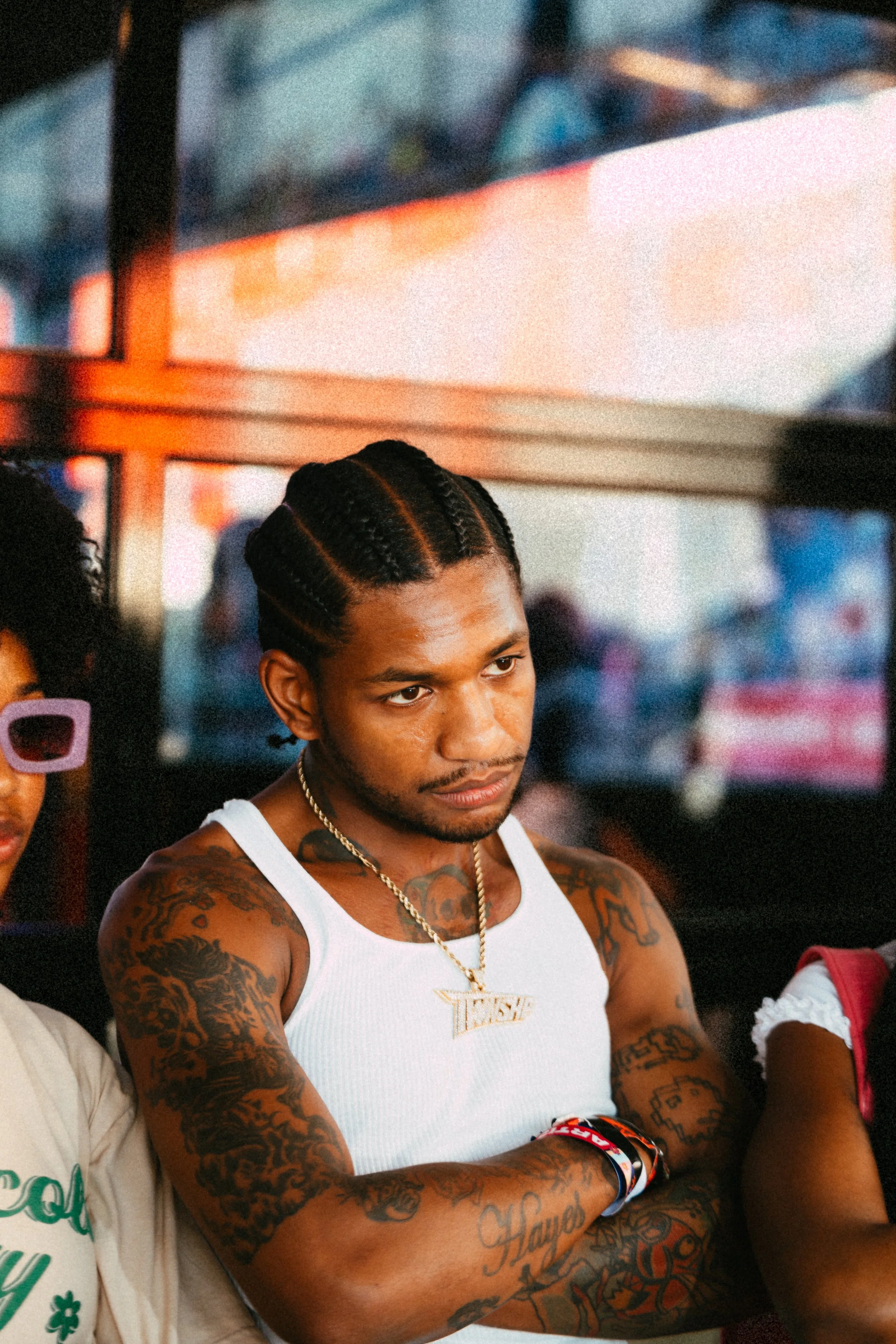 A man with braided hair and tattoos, wearing a white tank top and chain necklace, sitting with arms crossed, next to others.